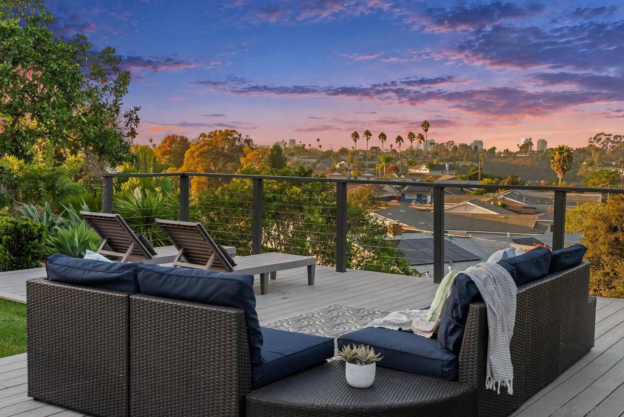 Rooftop deck with wicker lounge seating overlooks neighborhood rooftops beneath a colorful sunset sky.