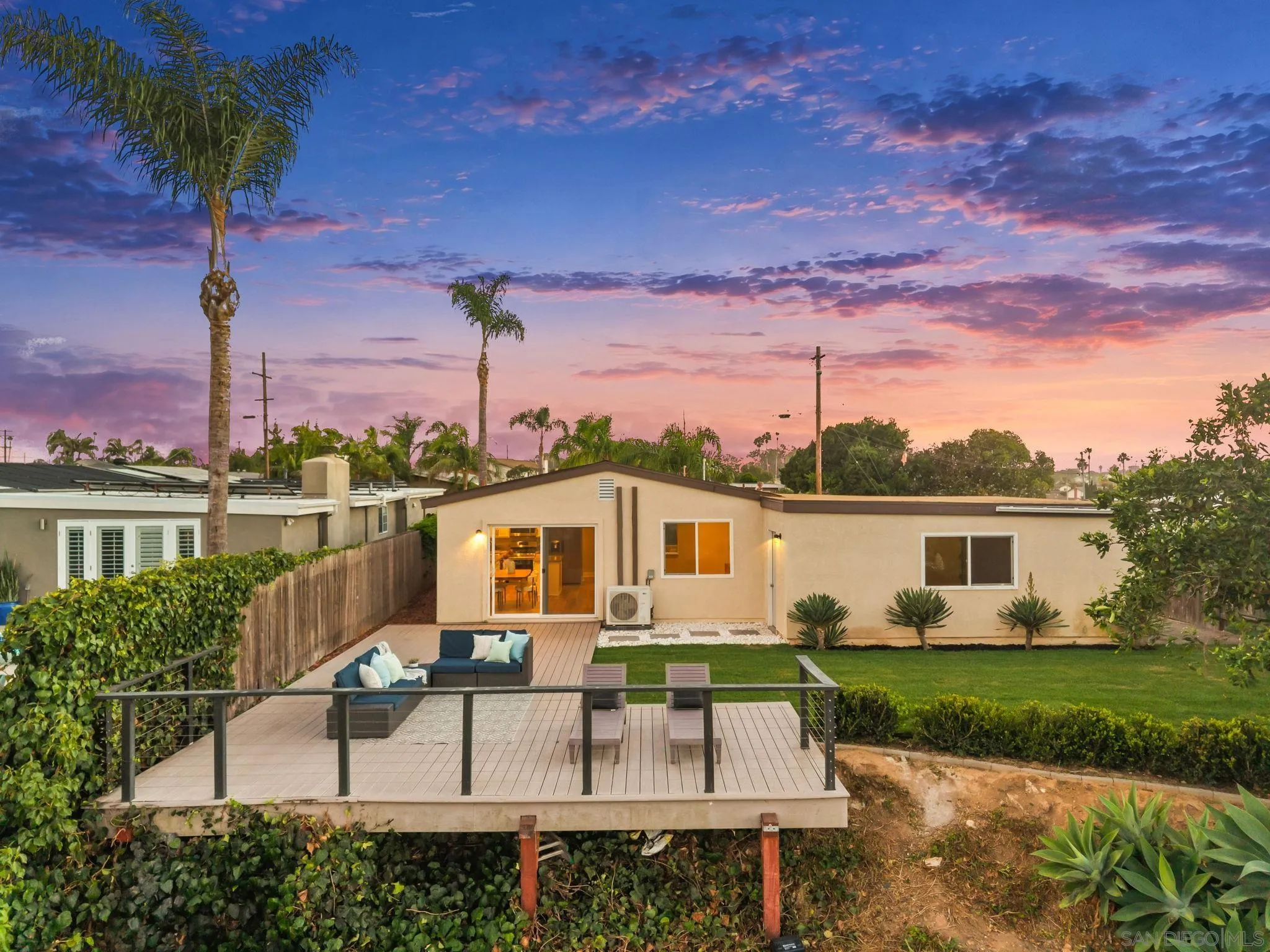 Home’s rear exterior at dusk, with a raised deck, outdoor seating, lawn, and palm trees.