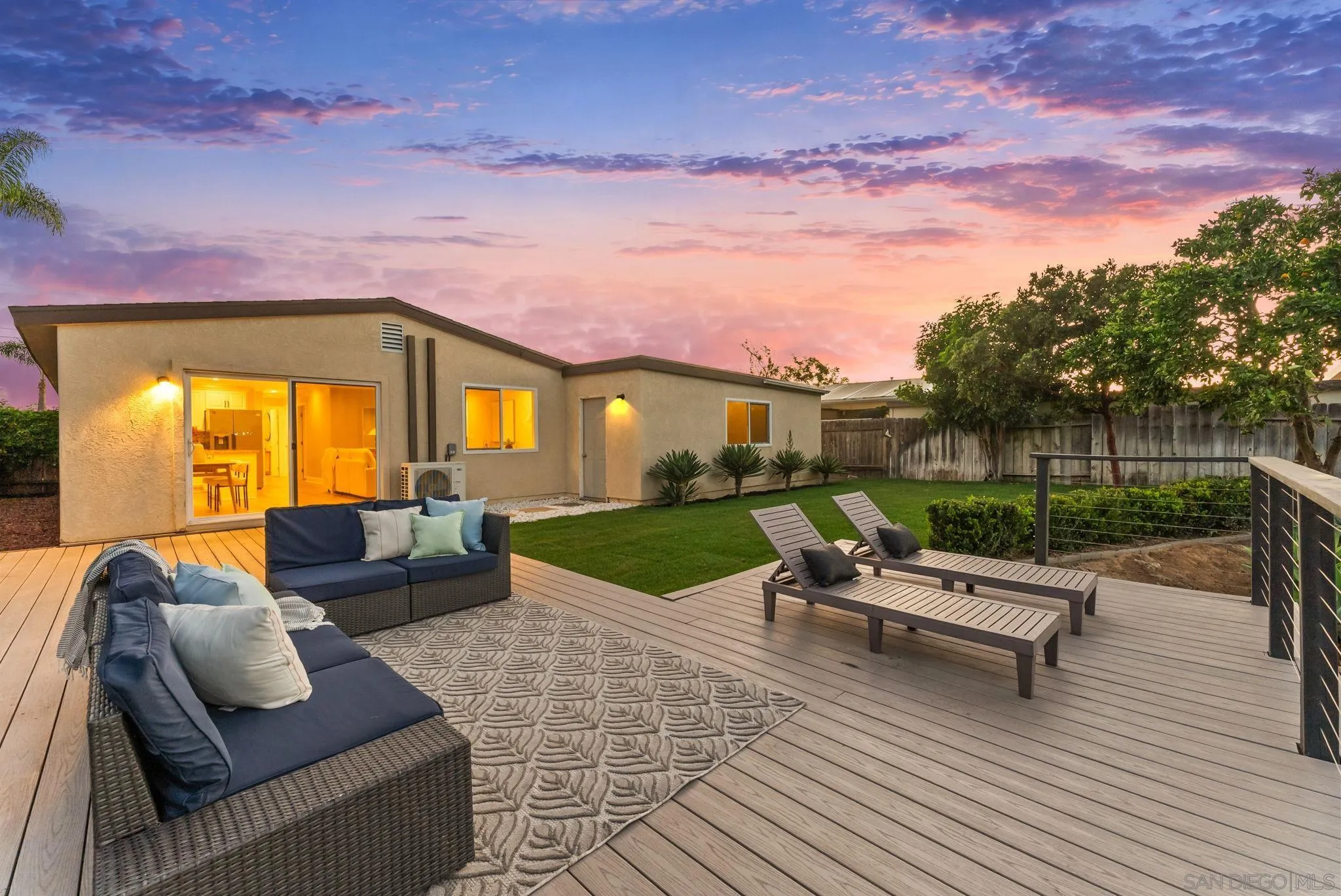 Backyard deck with lounge seating, lawn, and illuminated rear facade at dusk.