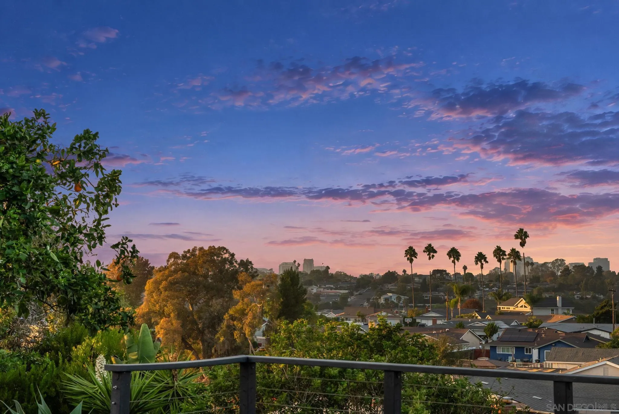 Neighborhood and distant city skyline at sunset, viewed over a garden terrace railing.
