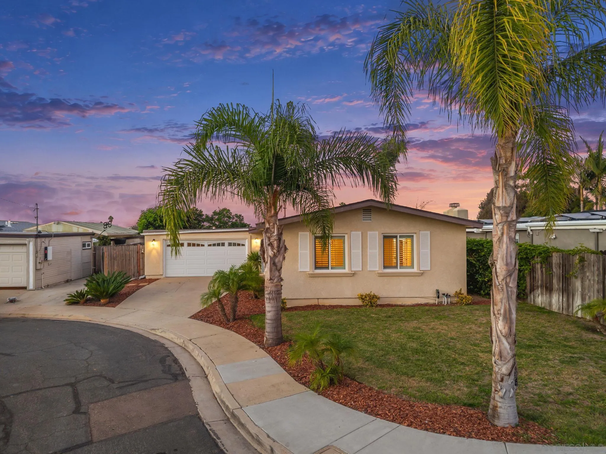 Single-story beige house with a double garage and palm trees at dusk