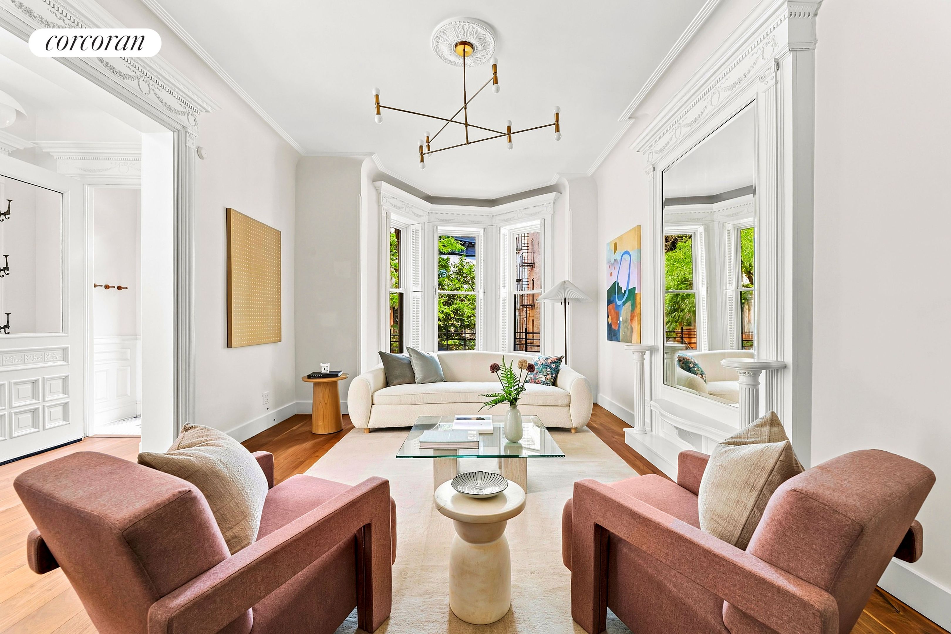 Staged living room with ornate moldings, leafy bay windows, a cream sofa, and rose-colored armchairs.