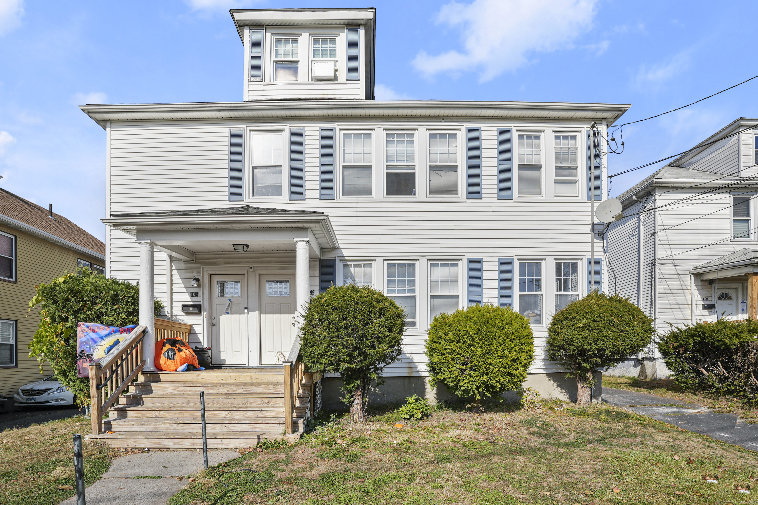White multi-unit house with blue shutters, raised wooden porch, and neatly trimmed front shrubs.