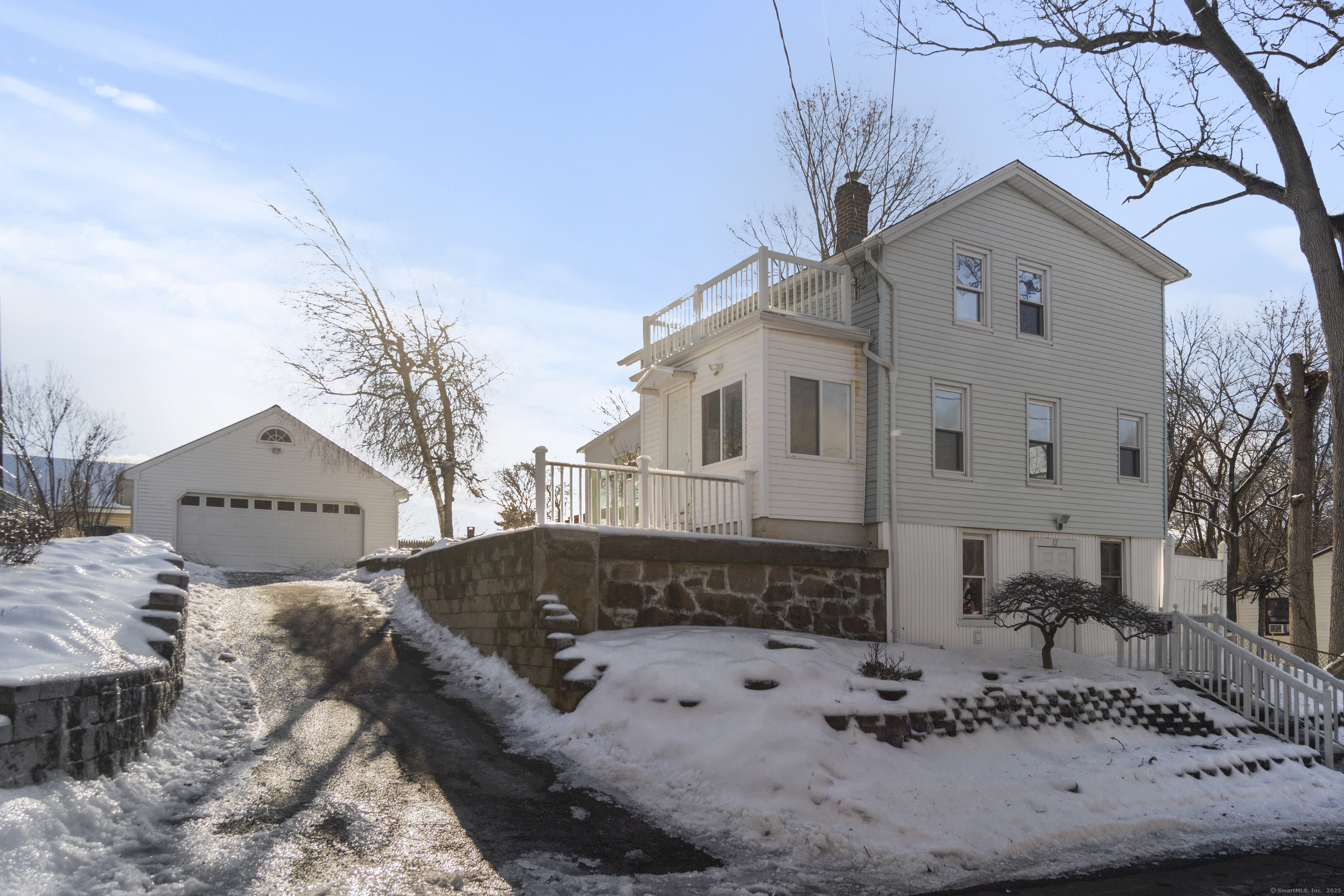 Two-story gray house with a raised deck, stone retaining wall, snowy yard, and detached garage.