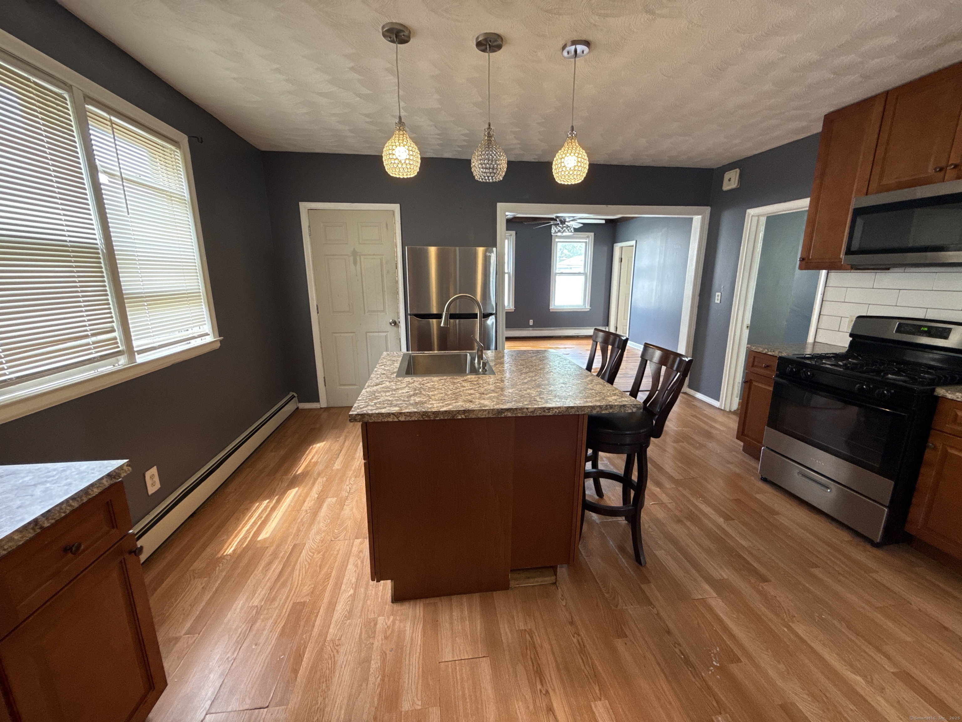 Kitchen with a large island, three pendant lights, stainless steel appliances, and an open doorway to another room.