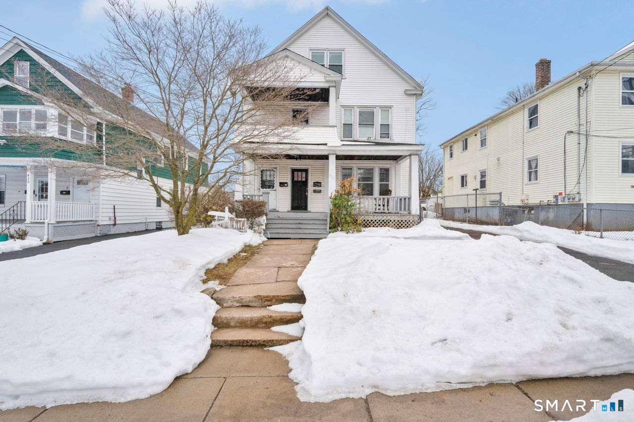 White multi-story house with a covered porch and snowbanks lining the front walkway.