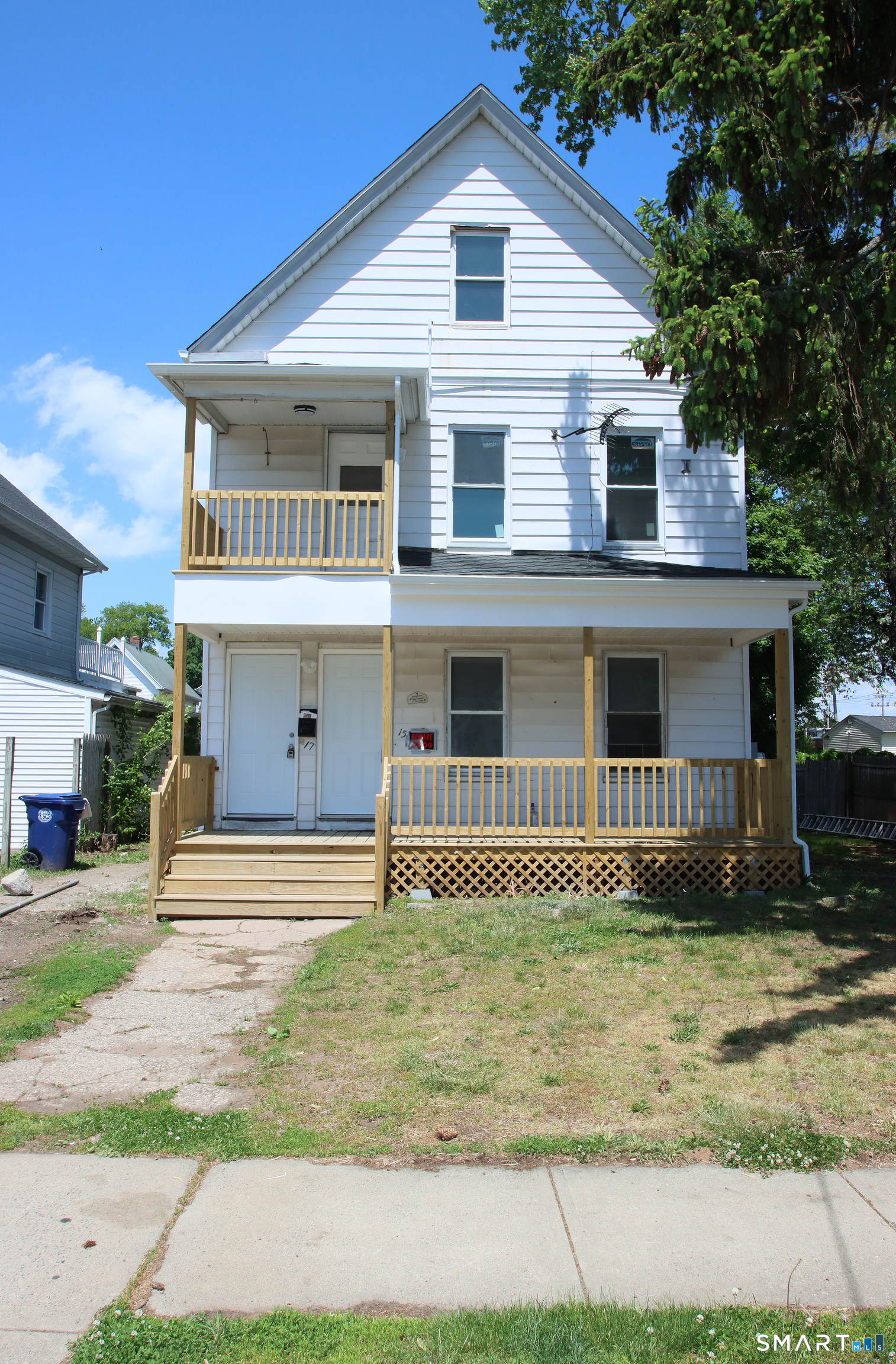 White-sided house with a steep front gable and two wooden porches, viewed from the street.