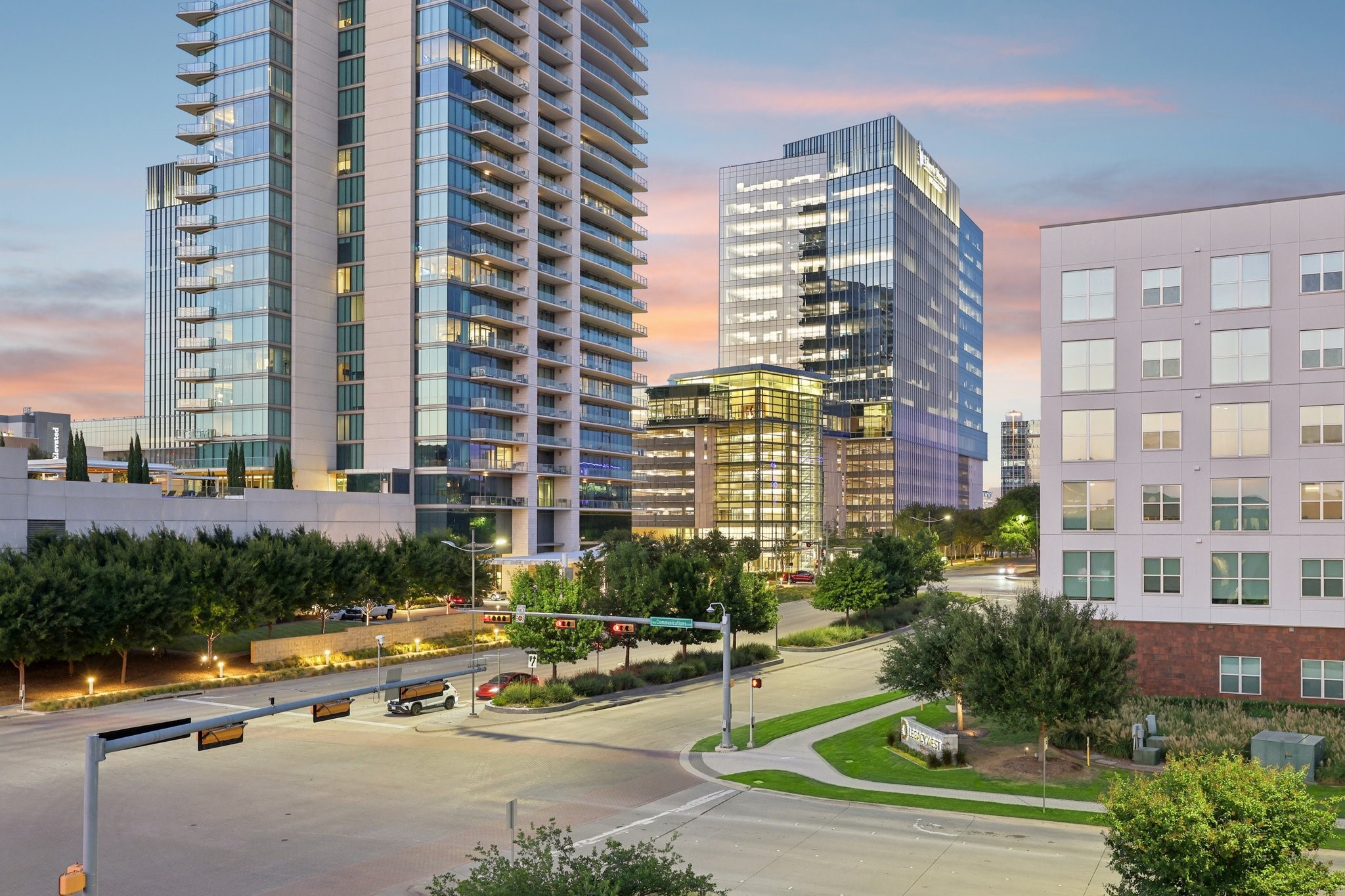 Modern high-rises and glass office towers at a landscaped intersection beneath a pink dusk sky.