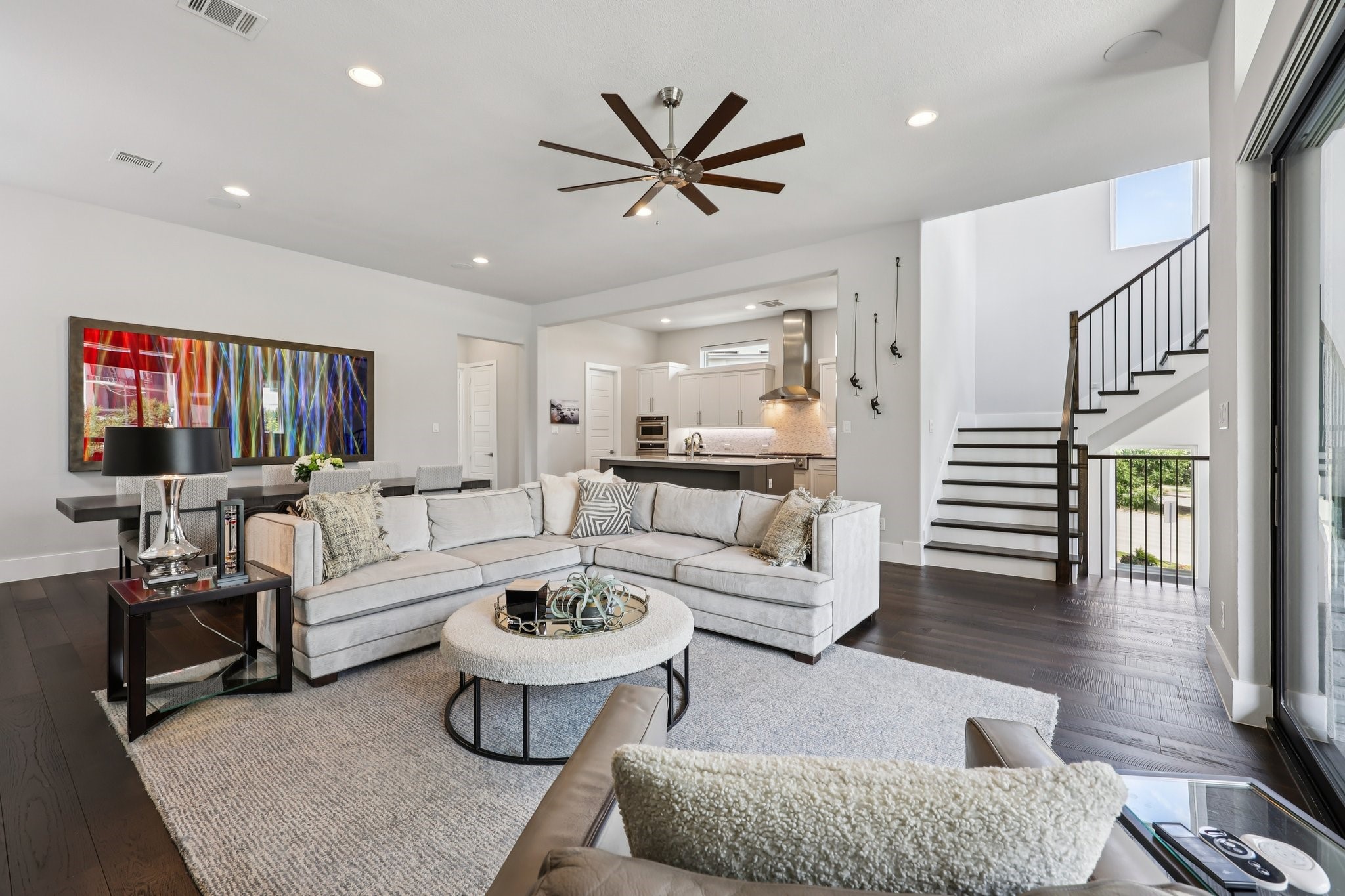 Open-plan living room with a light sectional, colorful wall art, kitchen, and staircase.