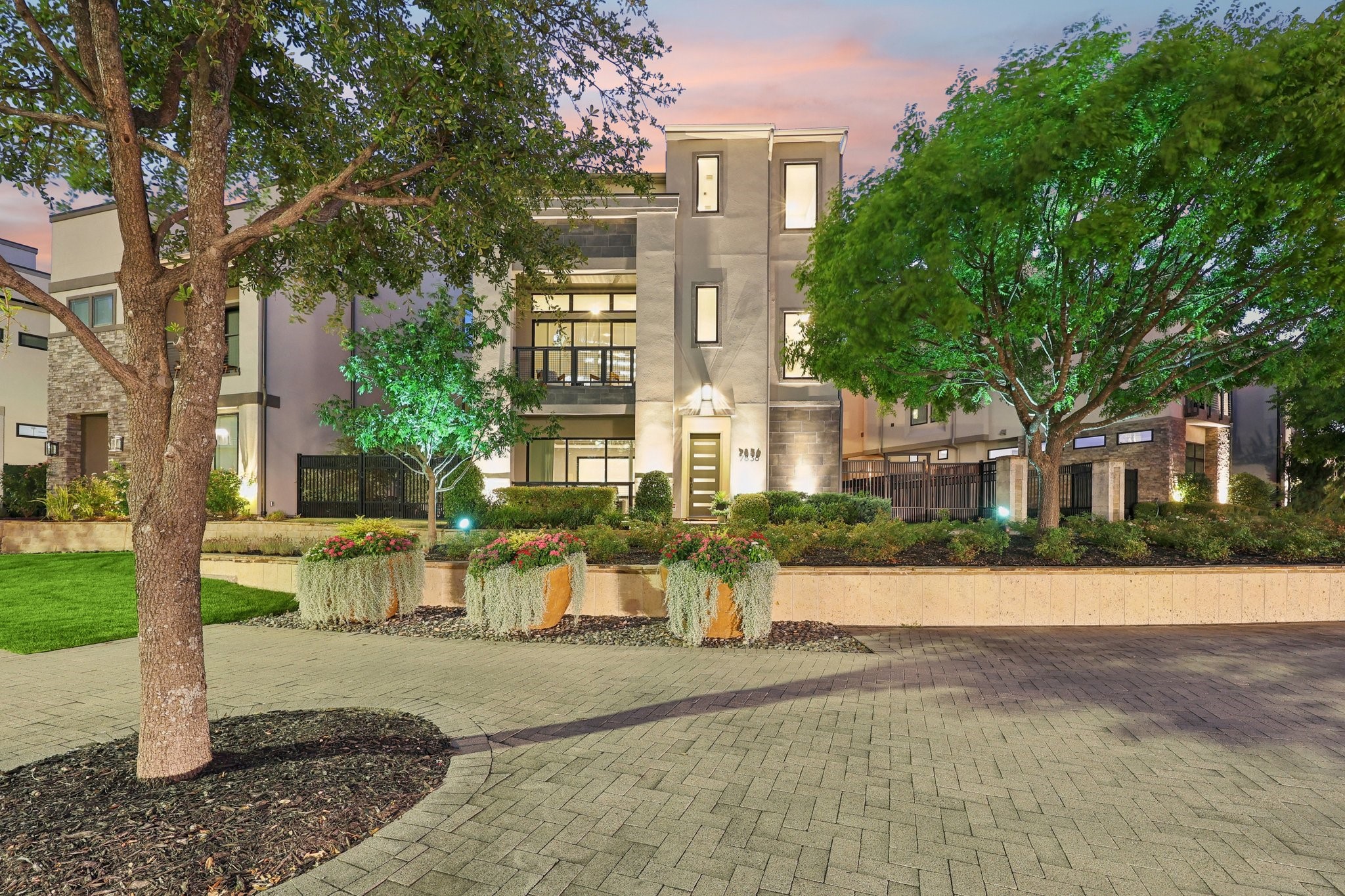 Modern multi-story home at dusk, framed by mature trees and flower planters along a paved courtyard.