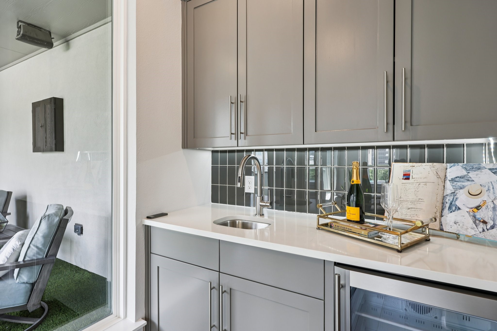 Gray wet bar with sink, glossy tile backsplash, and champagne setup beside a glass patio door.