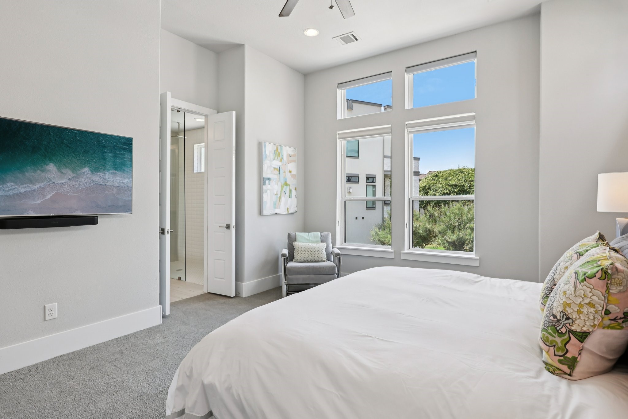 Bedroom with a large bed, tall windows, and a gray armchair beside the ensuite bathroom.