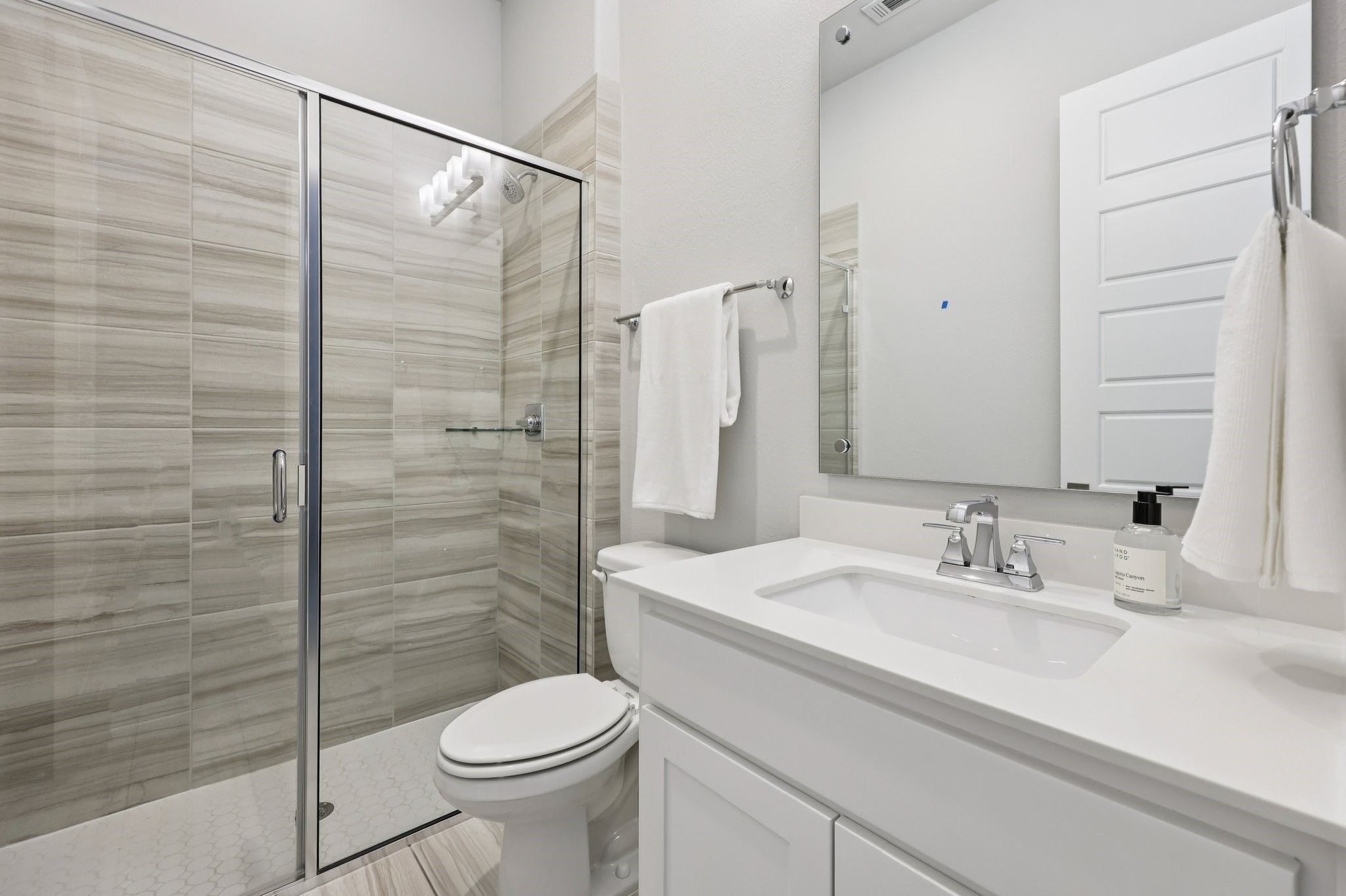 Bathroom with a glass-enclosed tiled shower, white toilet, and broad vanity beneath a large mirror.