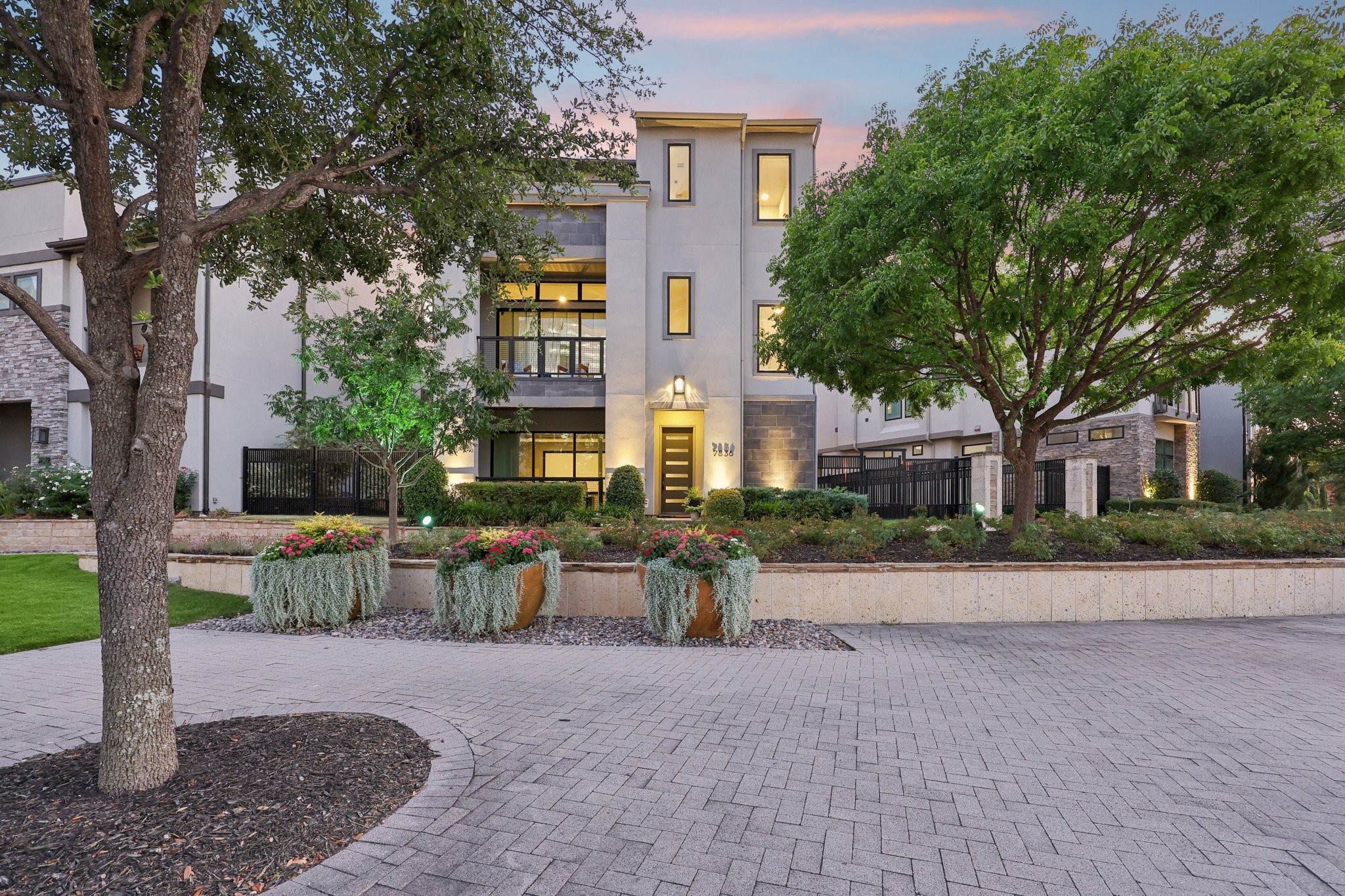 Modern multi-story home framed by mature trees, with illuminated windows and landscaped flower planters.