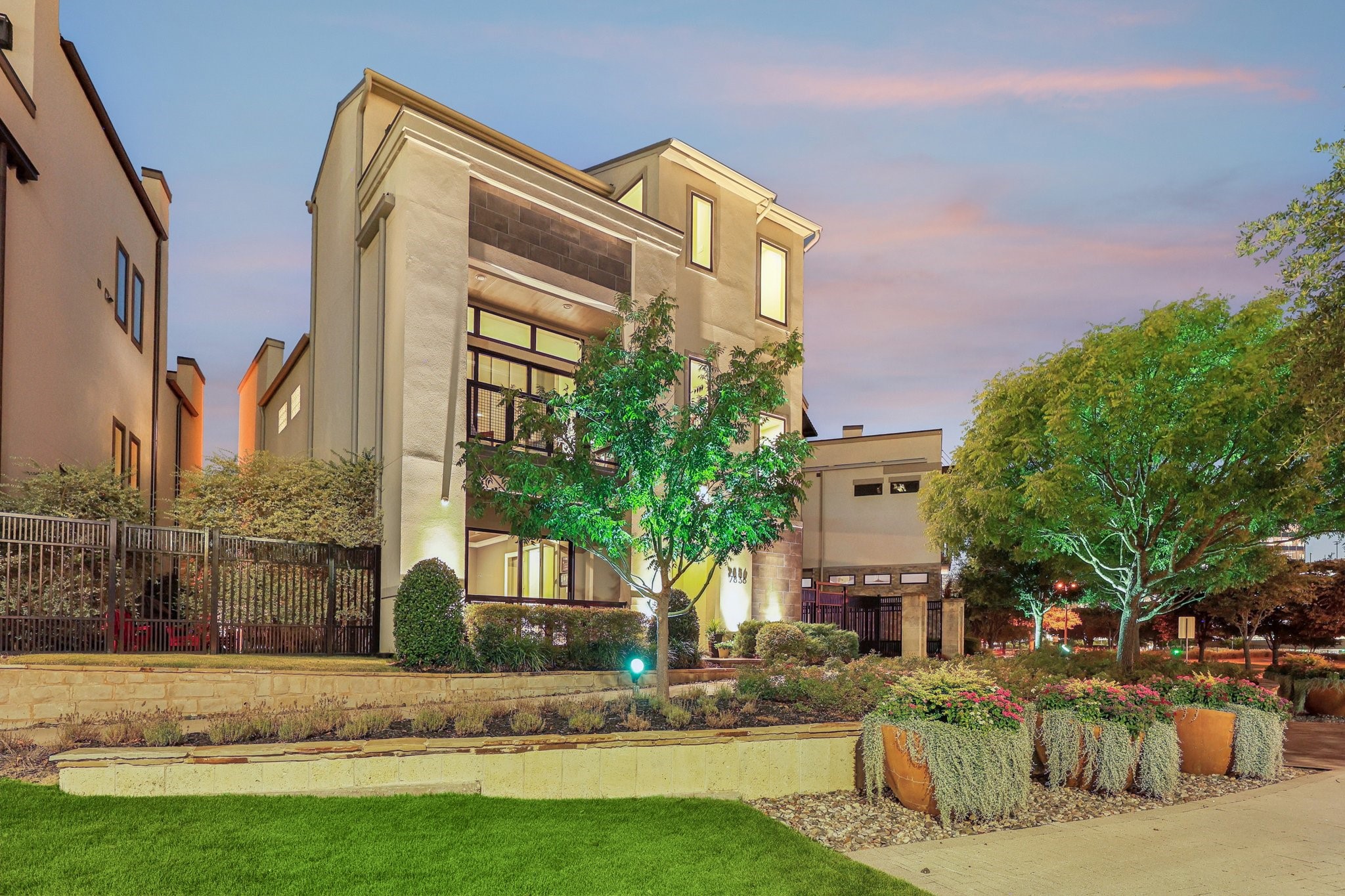 Multi-story home at dusk with illuminated windows, landscaped flower beds, and mature trees.