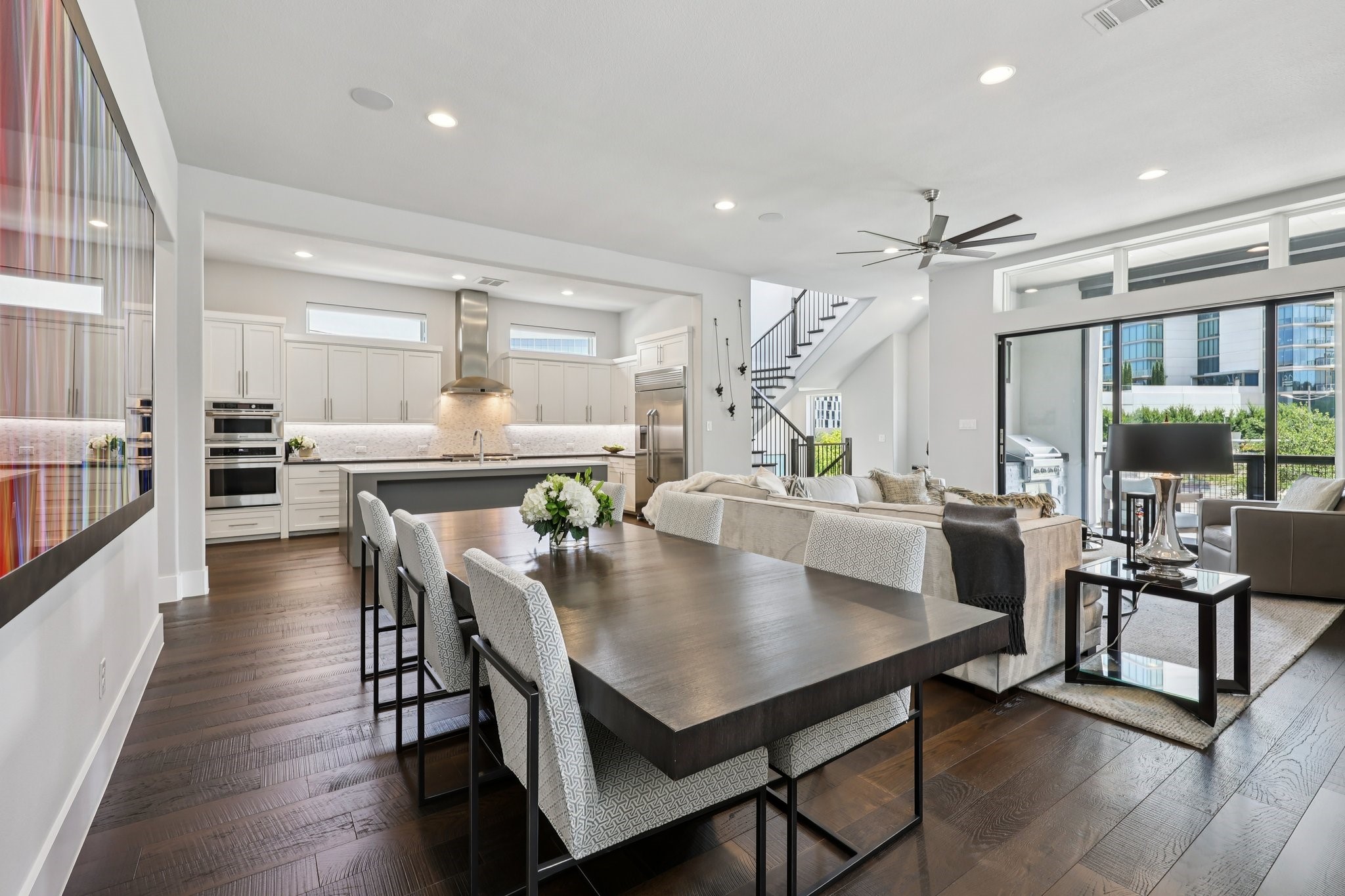 Open-plan kitchen and living room with a large dining table and balcony doors overlooking modern buildings.