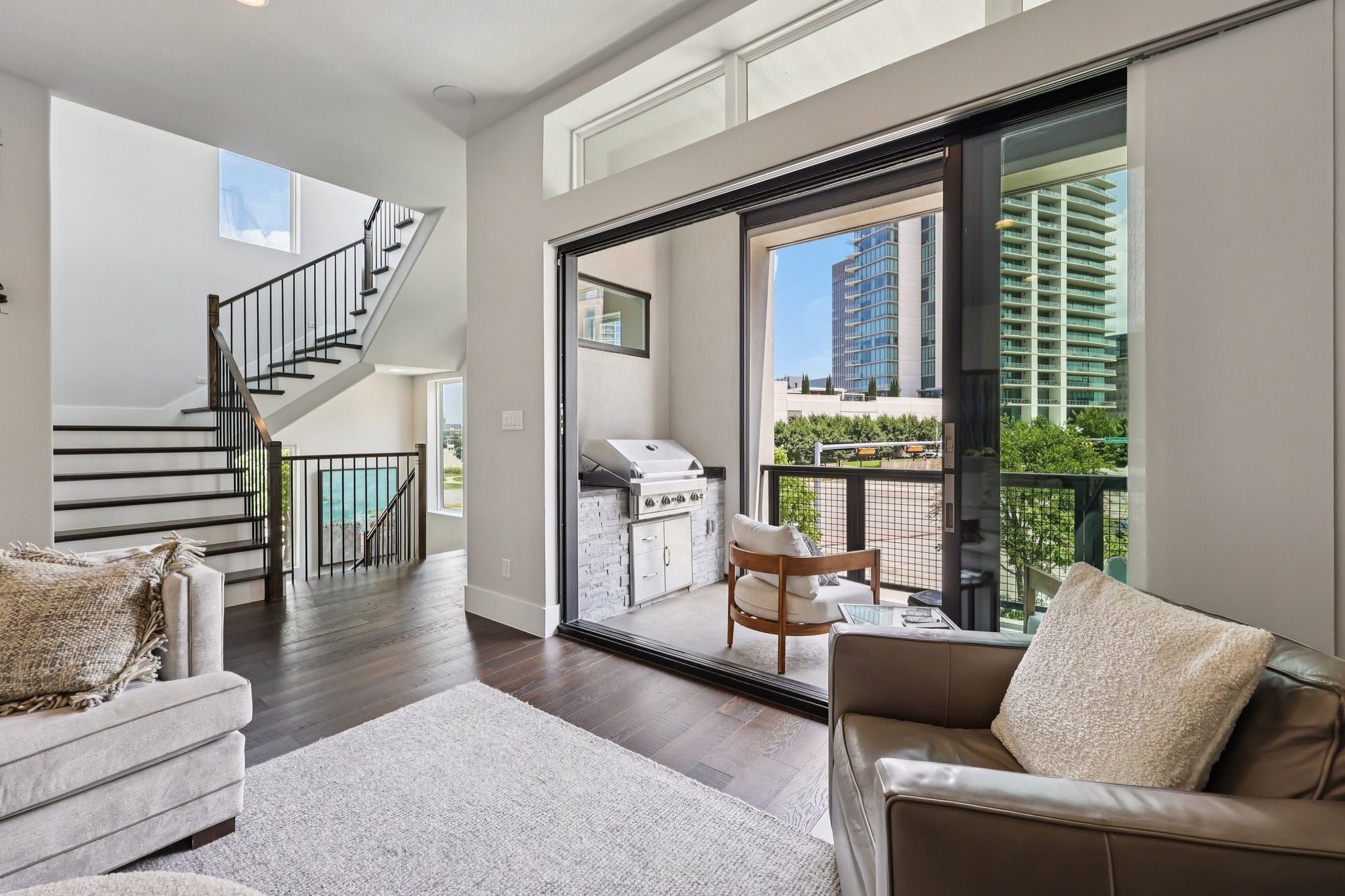 Modern living room with a dramatic staircase and sliding doors opening to a grill-equipped balcony overlooking city buildings.