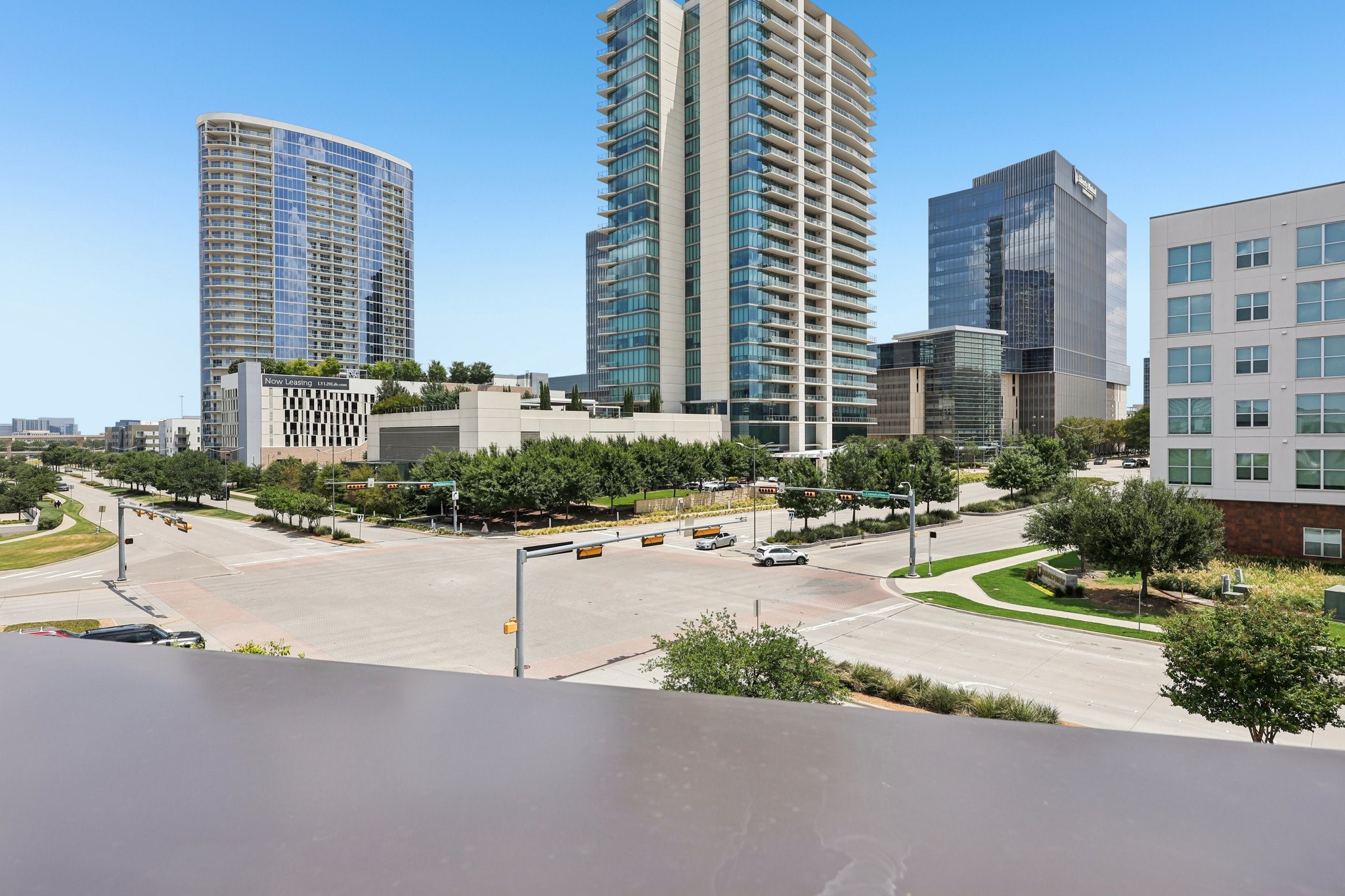 Urban high-rises and a landscaped intersection viewed from above, with a rooftop edge in the foreground.
