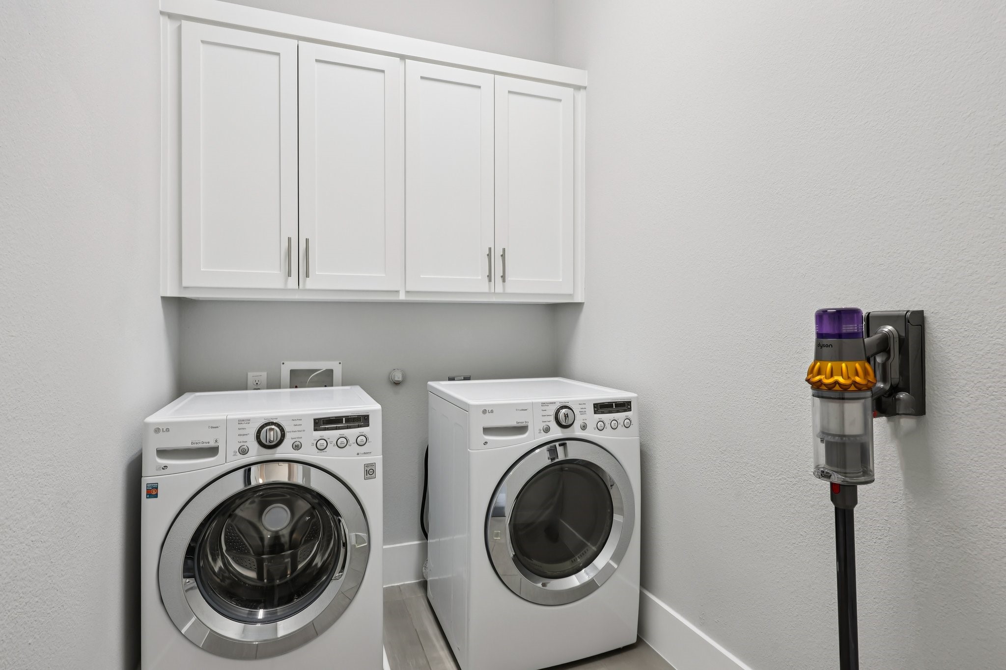 Laundry room with white cabinets, front-loading washer and dryer, and a mounted cordless vacuum.