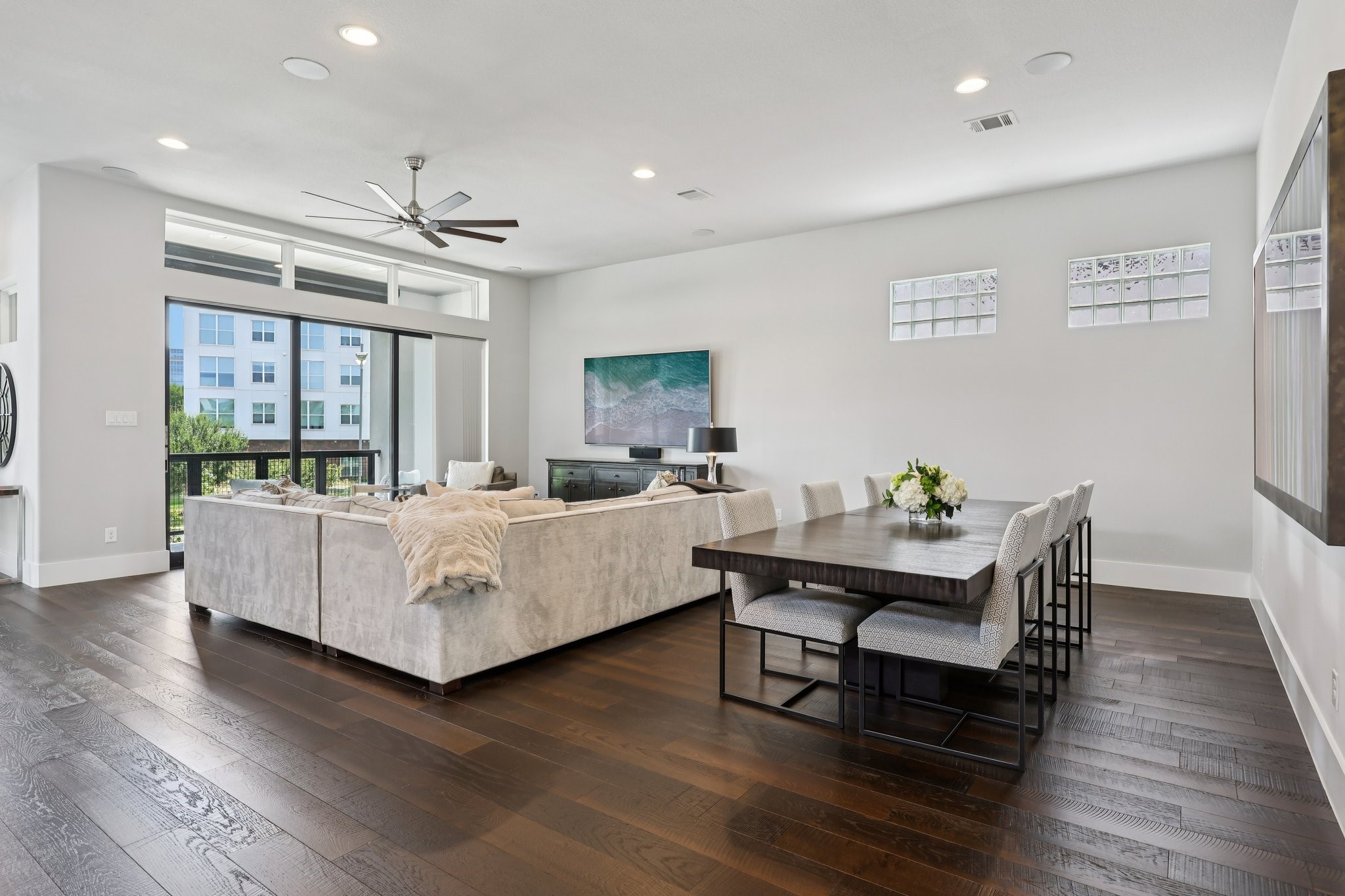 Open-plan living and dining room with a light sectional, dark hardwood floors, and balcony doors.