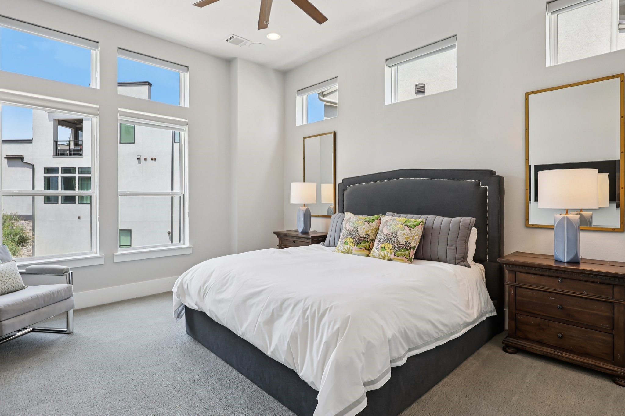 Bright staged bedroom with a large bed, dark headboard, and tall windows.