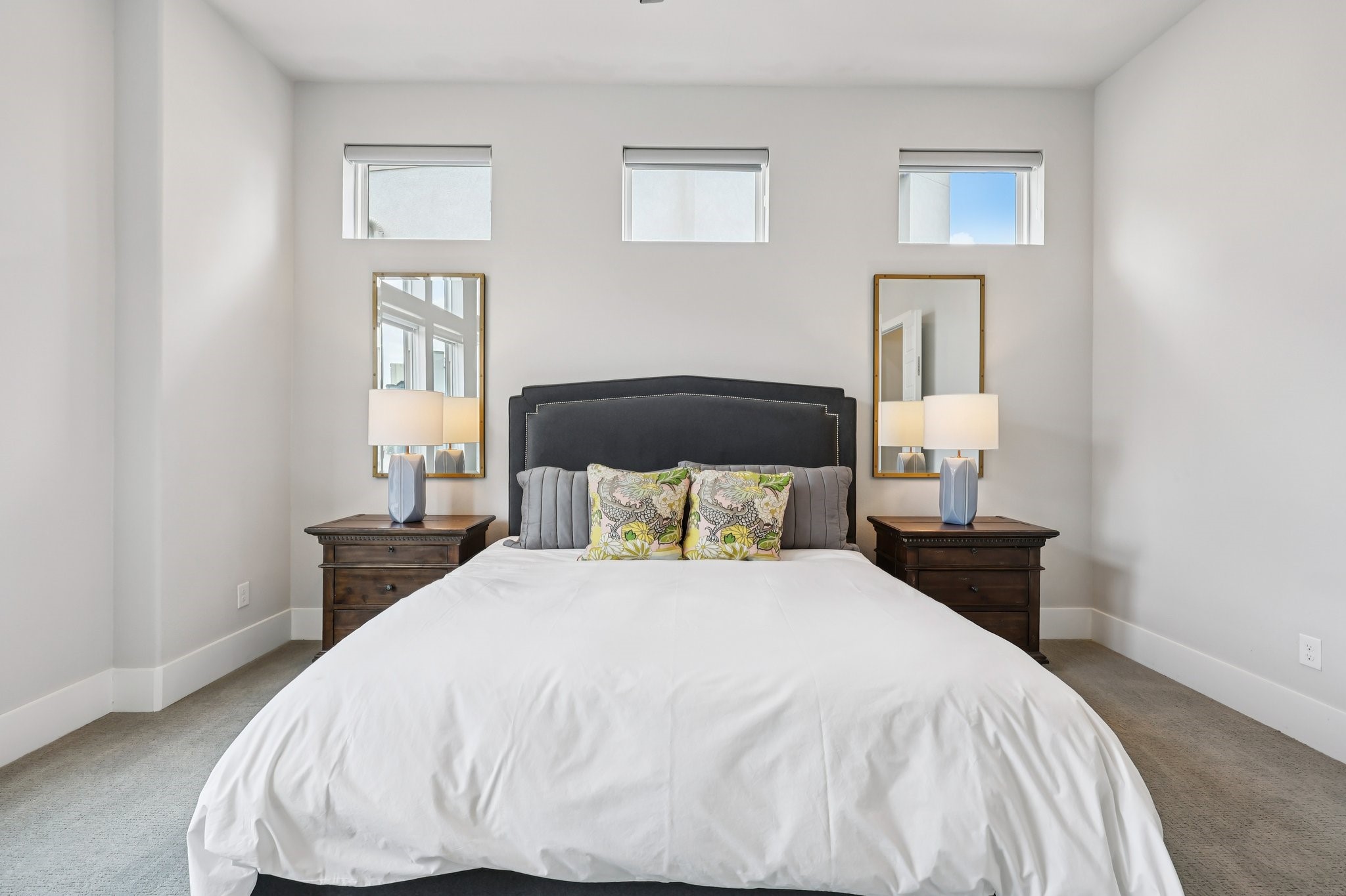 Staged bedroom with a large white bed, dark headboard, matching nightstands, and three high windows.