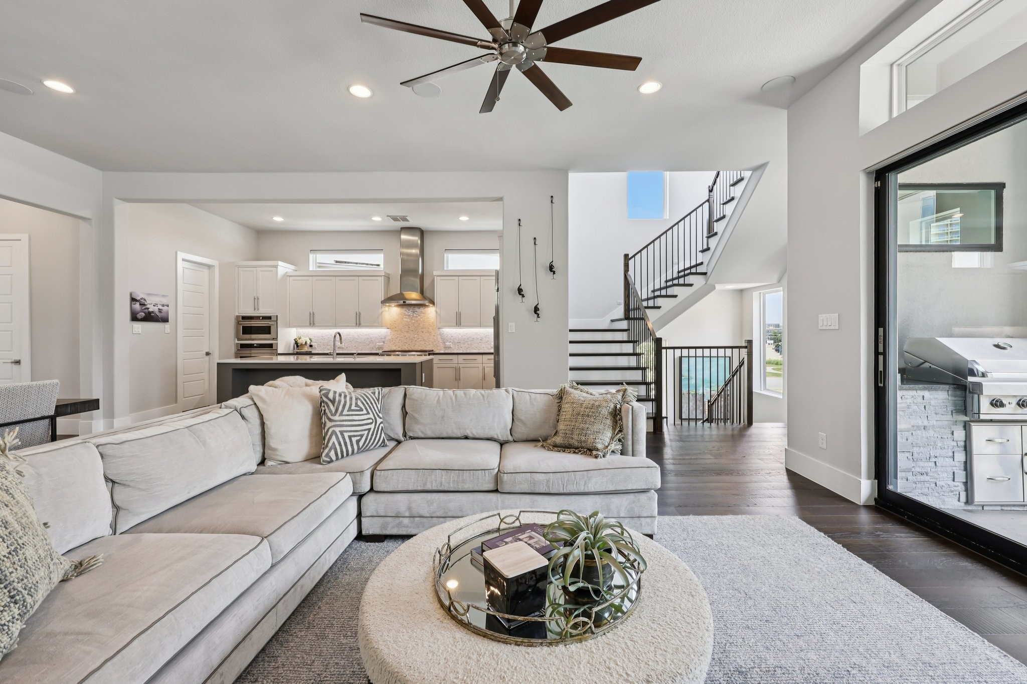 Open-plan living room with a large sectional, modern kitchen, black-railed staircase, and grill visible beyond sliding doors.