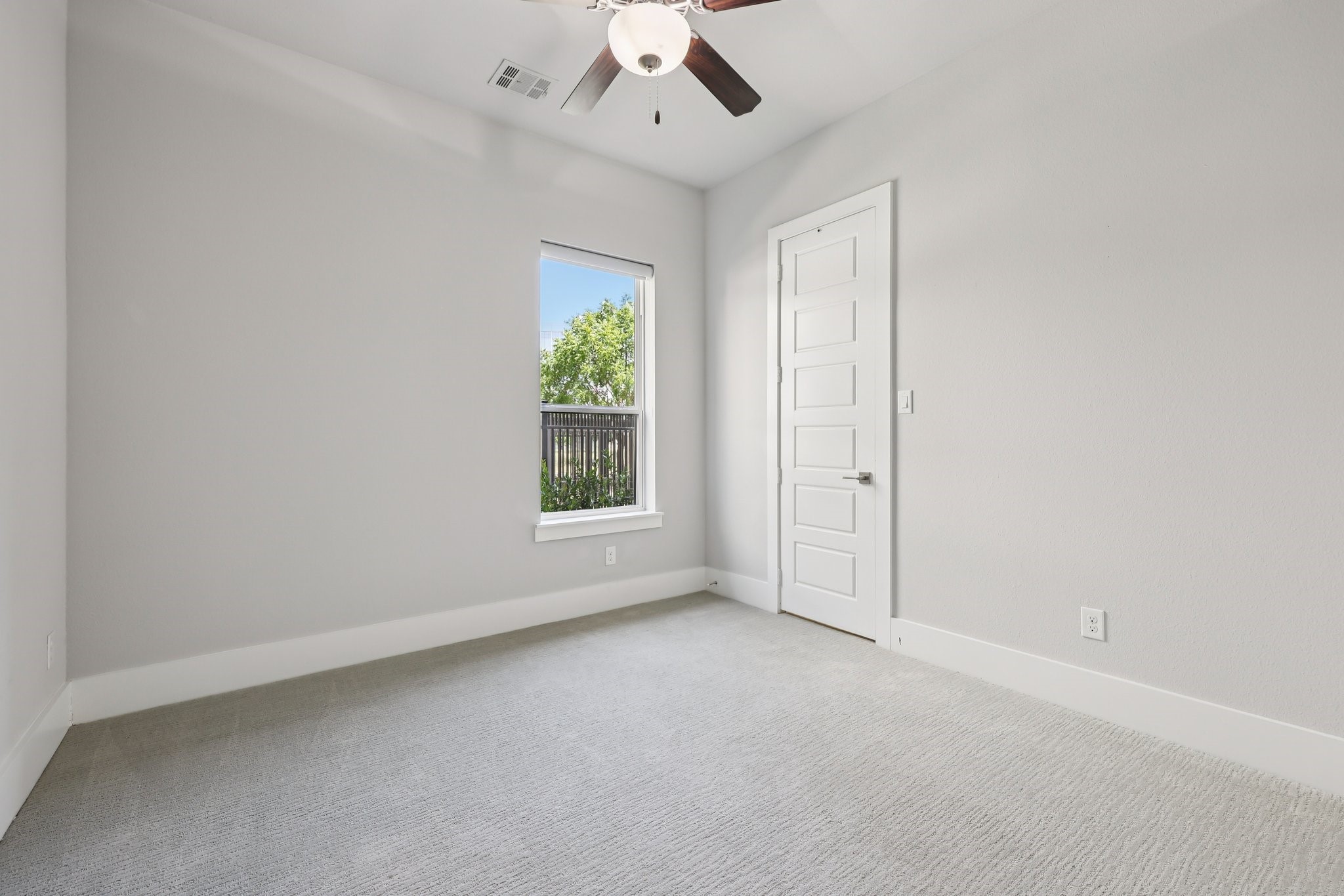 Empty carpeted room with a ceiling fan, a window overlooking a tree, and a white paneled door.