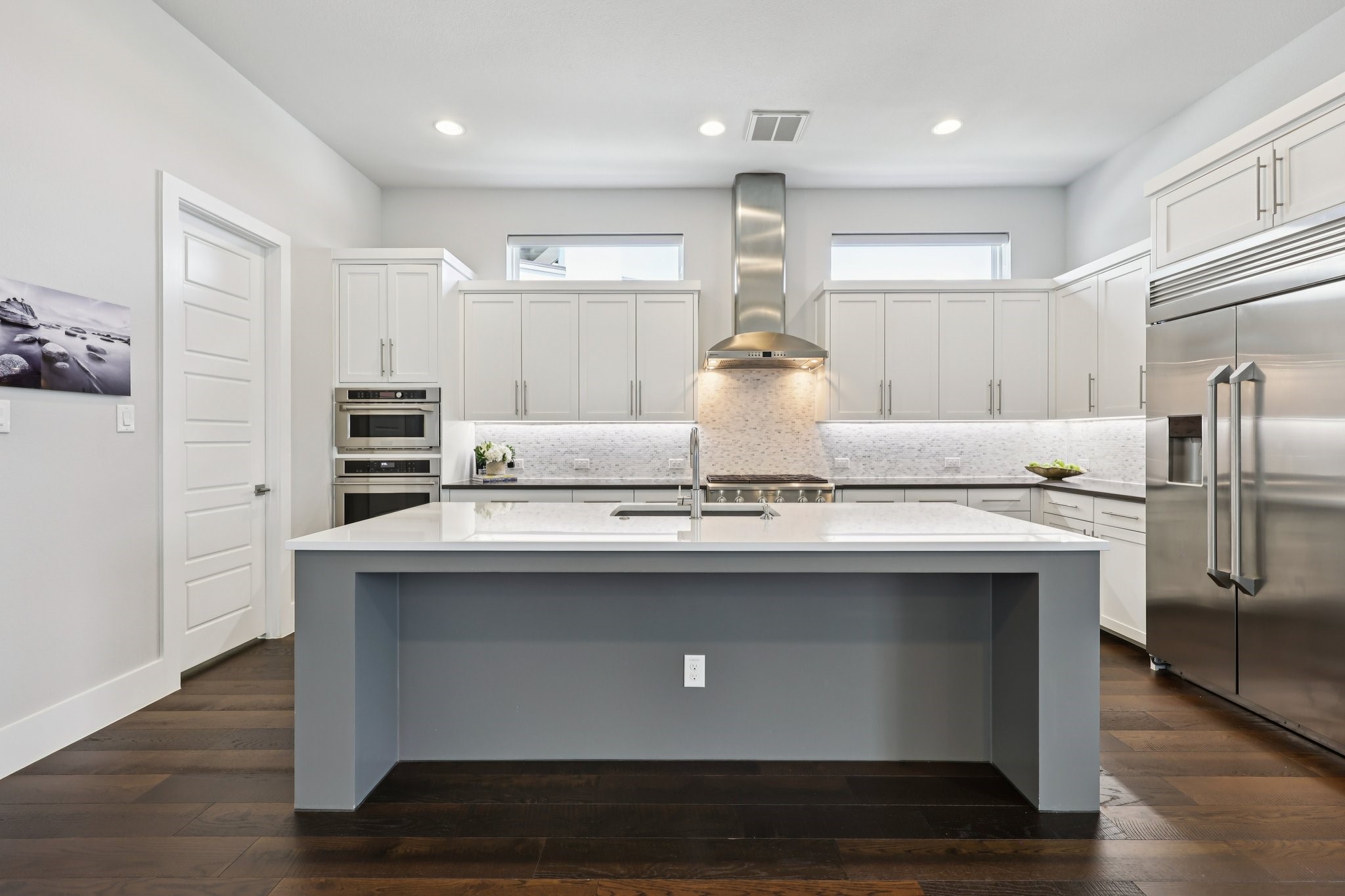 Modern white kitchen with a large gray island, stainless steel appliances, and dark wood flooring.
