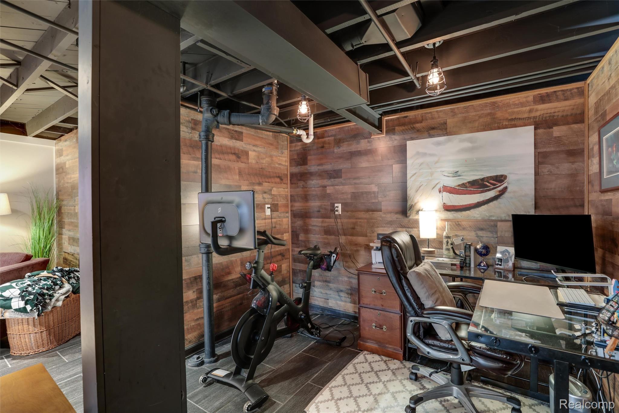 Home office with a glass desk, leather chair, exercise bike, and rustic wood-plank walls.