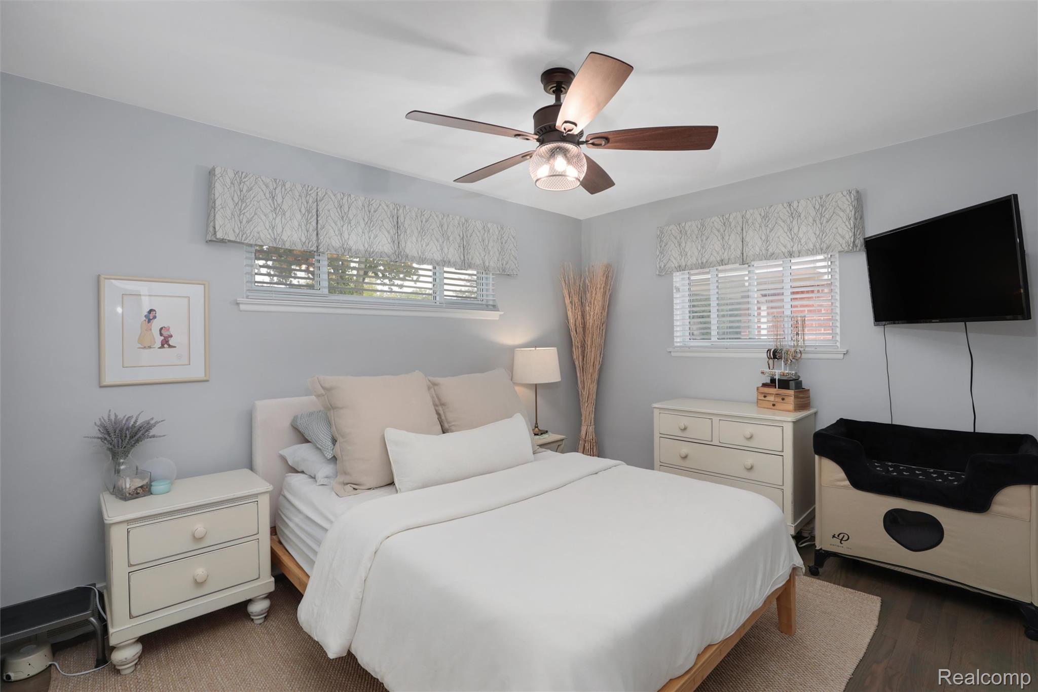 Furnished bedroom with a white bed, ceiling fan, two windows, and wall-mounted television.