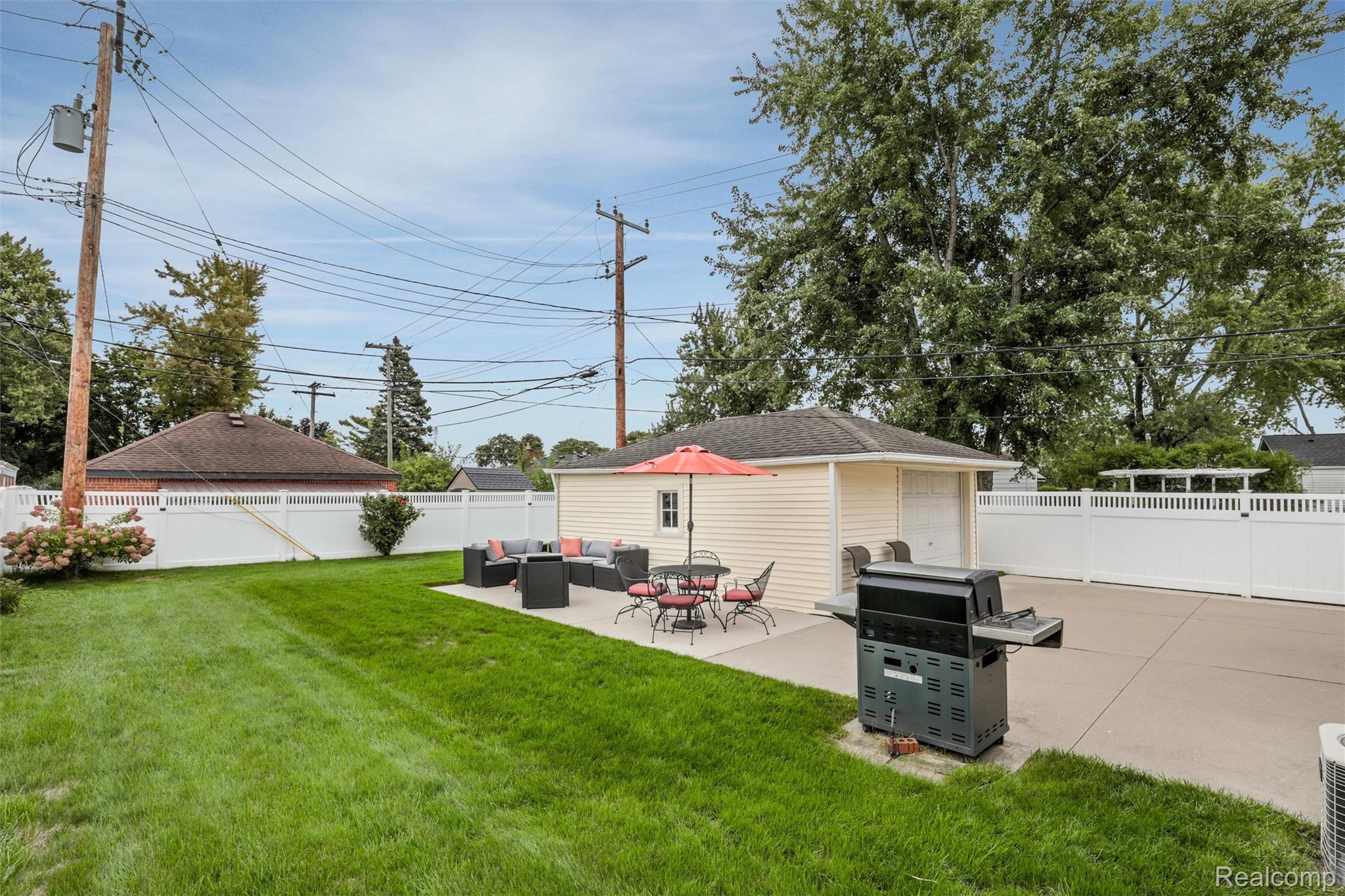 Backyard patio with lounge seating, dining set, coral umbrella, and grill beside a detached garage.