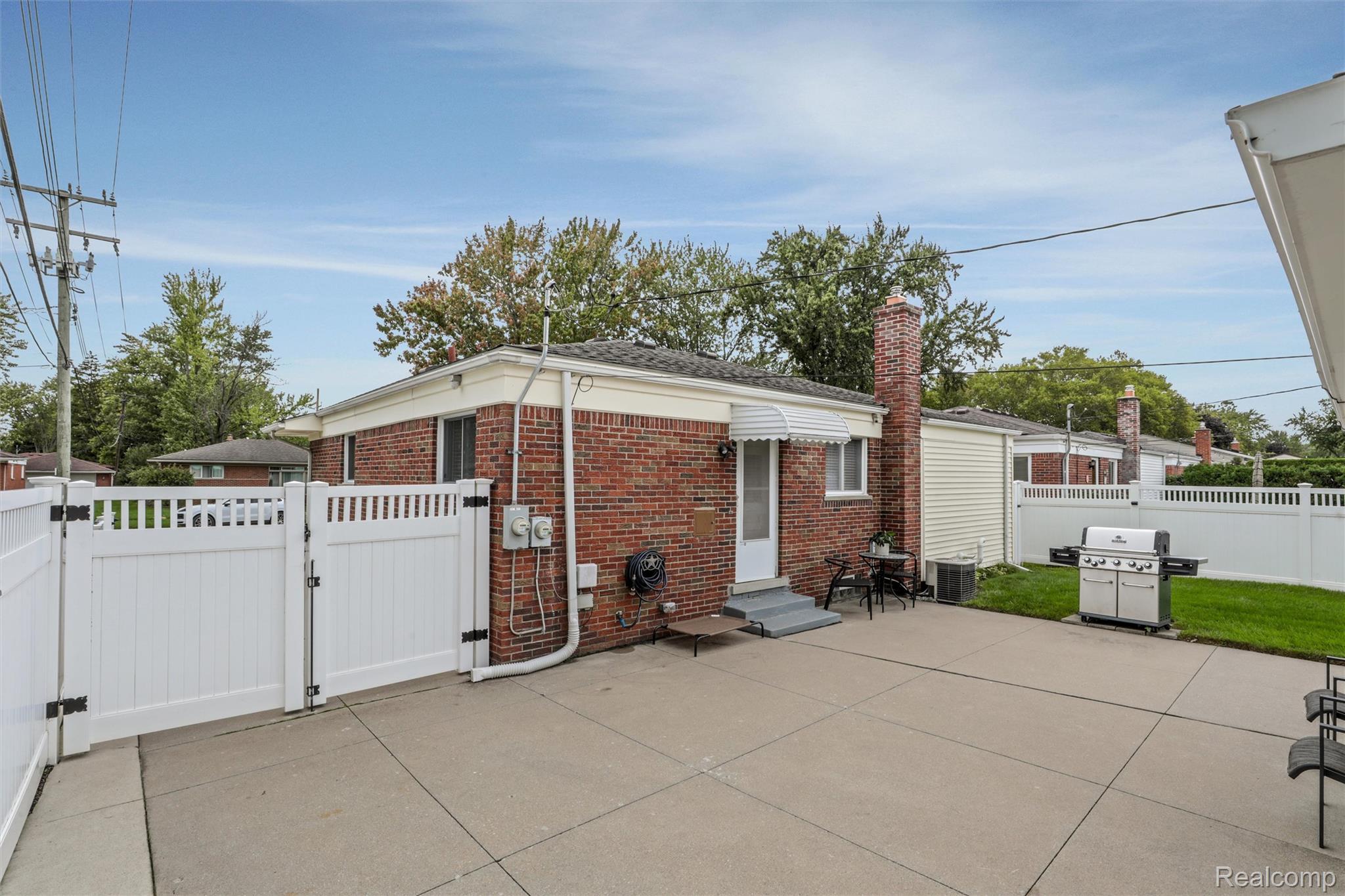 Red-brick house and fenced concrete patio with a white gate, grill, and outdoor seating.