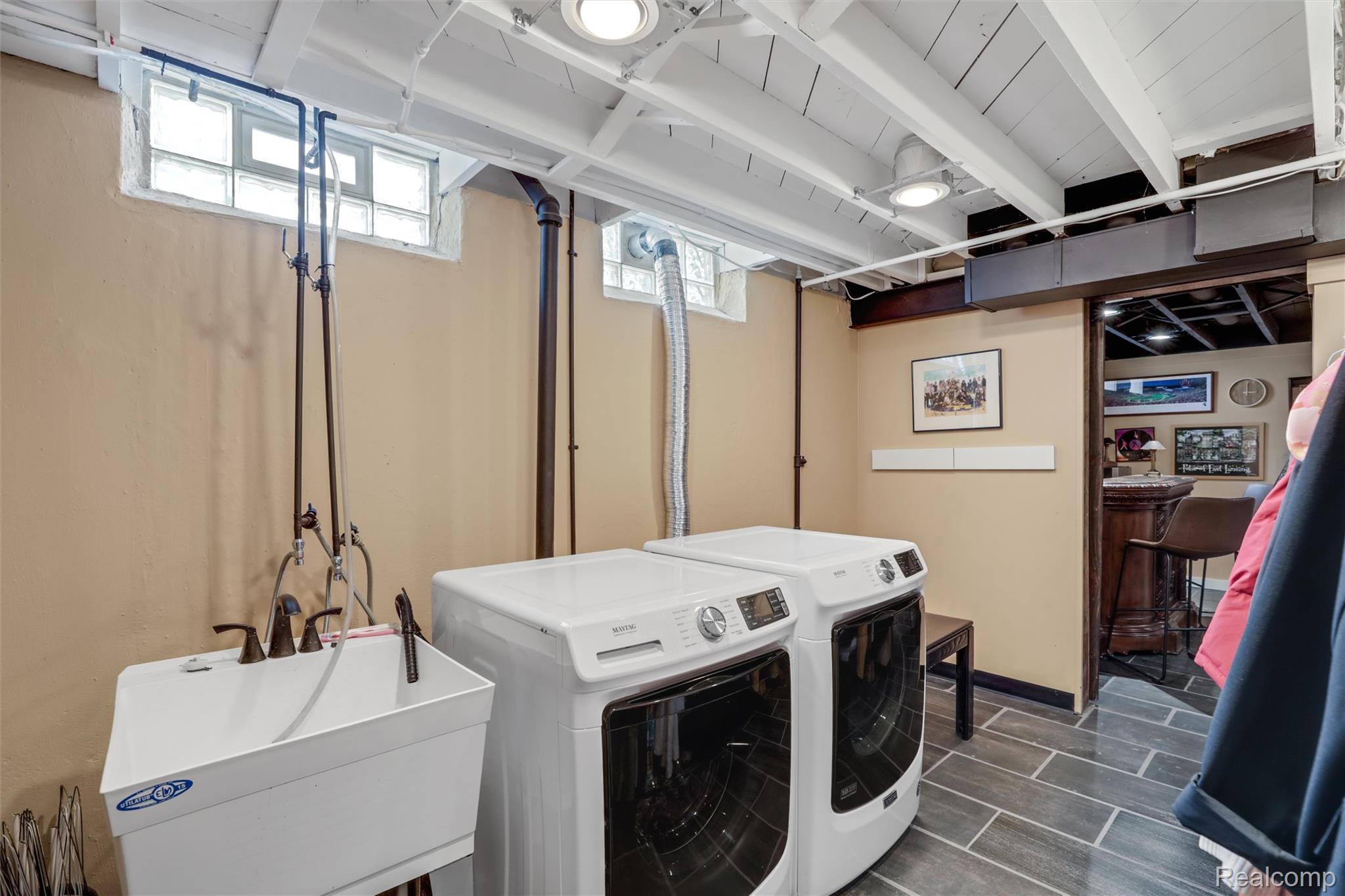 Basement laundry room with two white front-loading machines, a utility sink, and exposed ceiling beams.