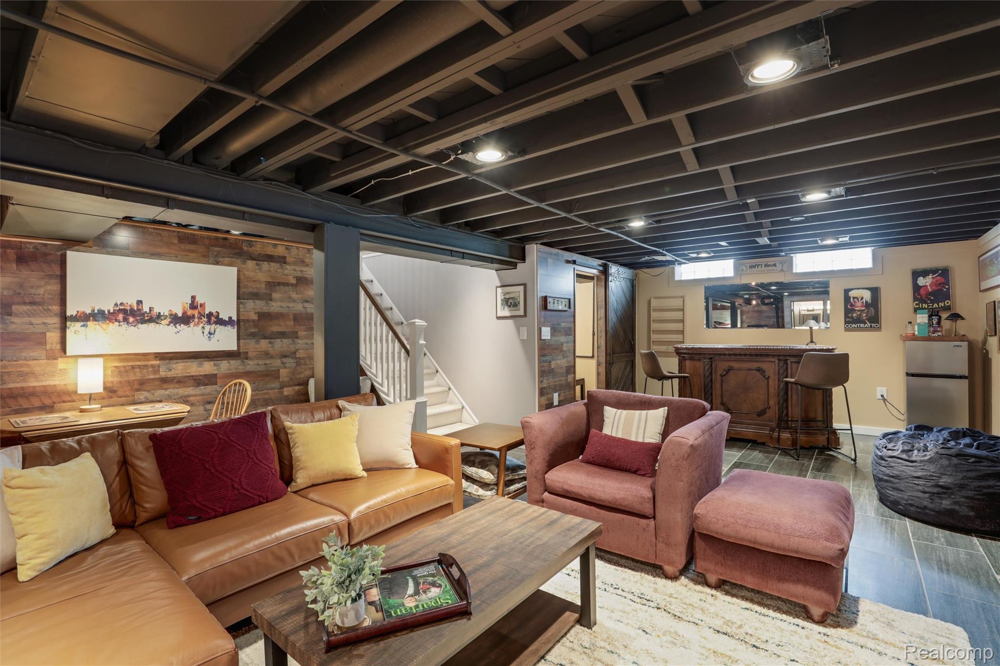 Basement recreation room with a leather sectional, pink armchair, exposed ceiling beams, and a built-in bar.