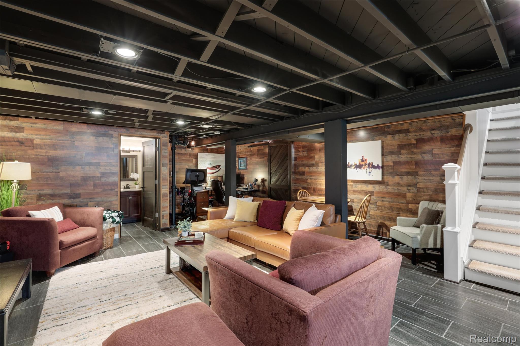 Finished basement living room with reclaimed wood walls, exposed ceiling beams, and a carpeted staircase.