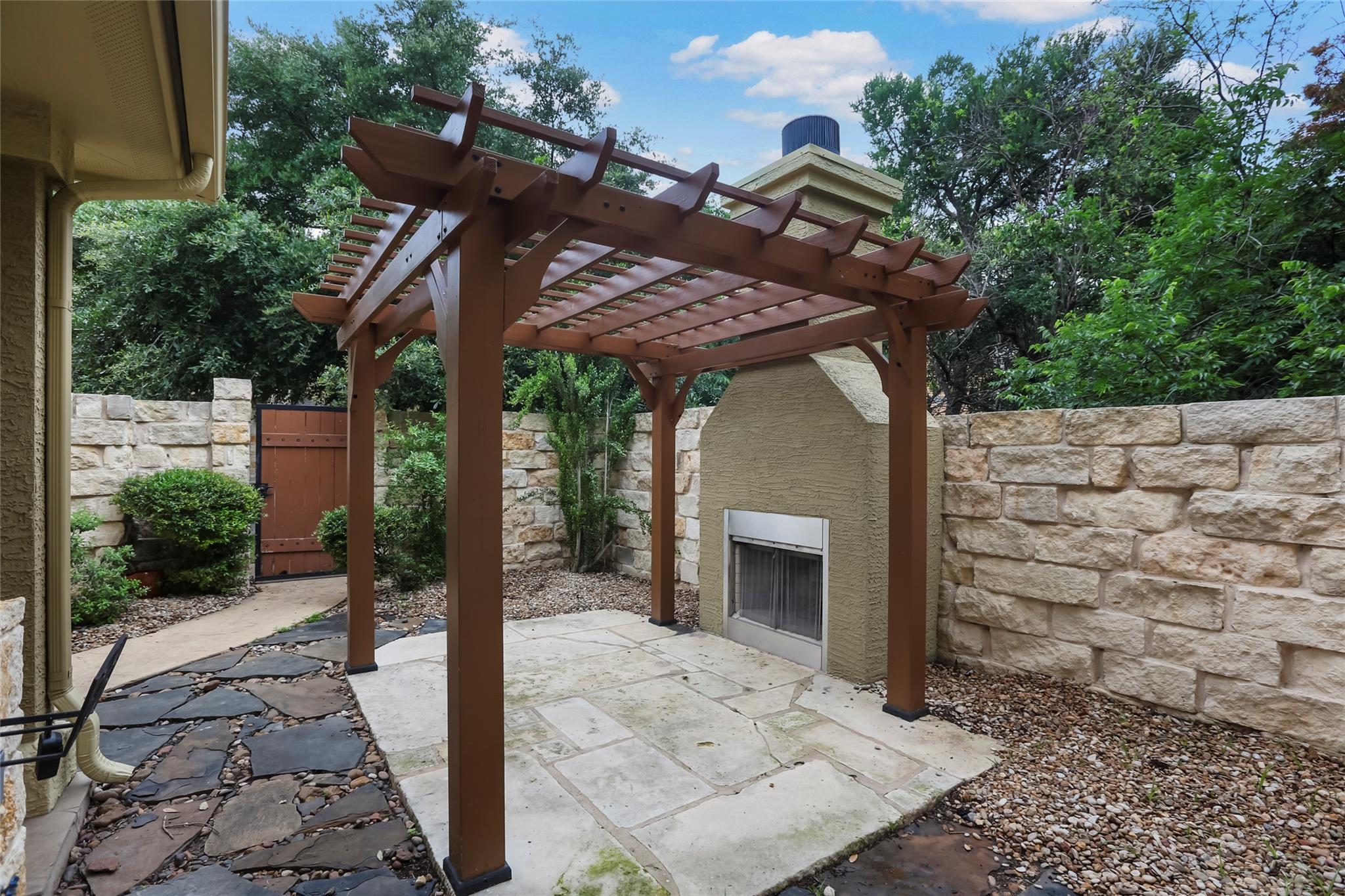 Stone patio beneath a wooden pergola, with a built-in fireplace and landscaped stone walls.
