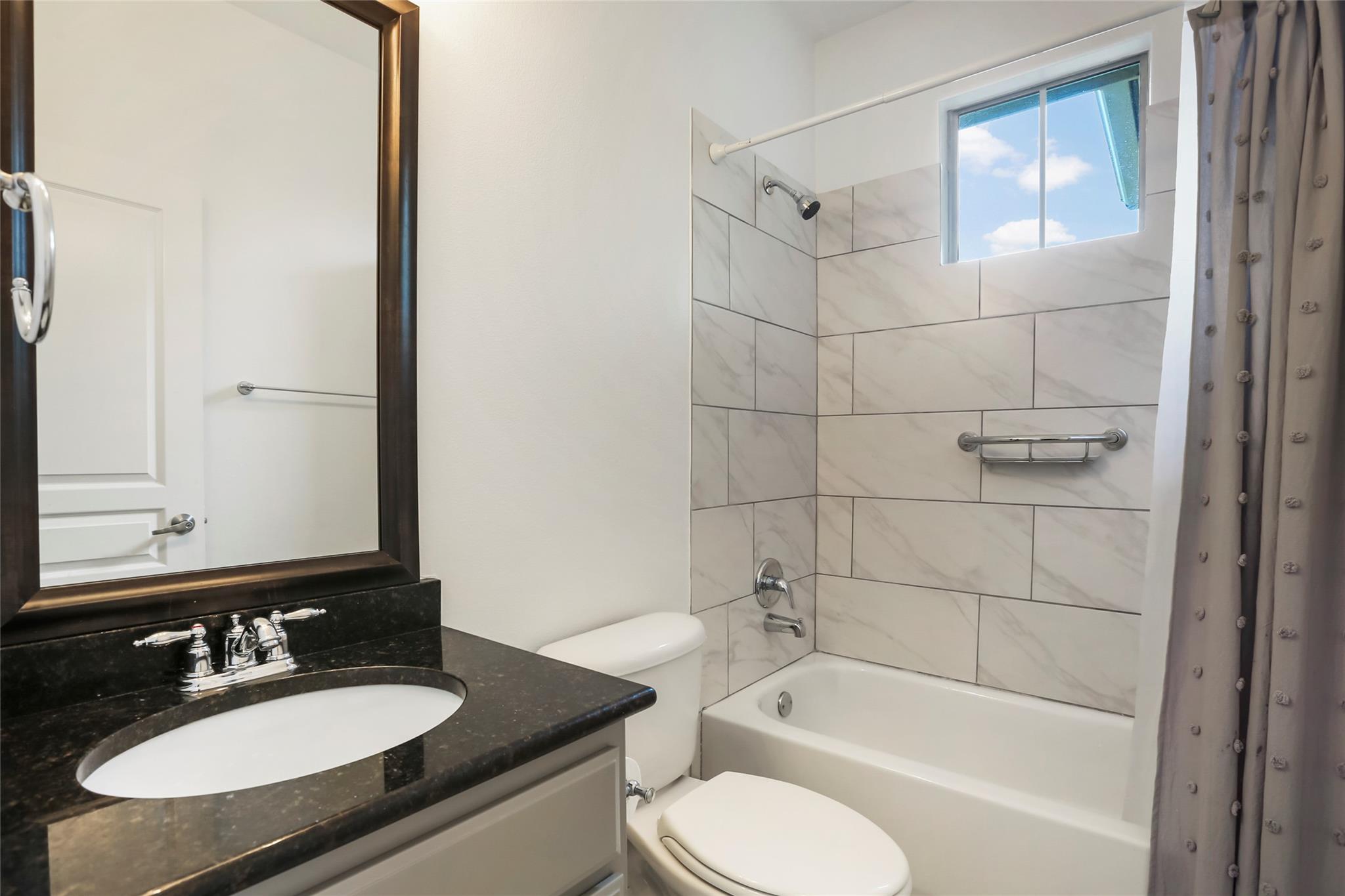 Bathroom with a tiled tub surround, high window, toilet, and dark stone vanity.