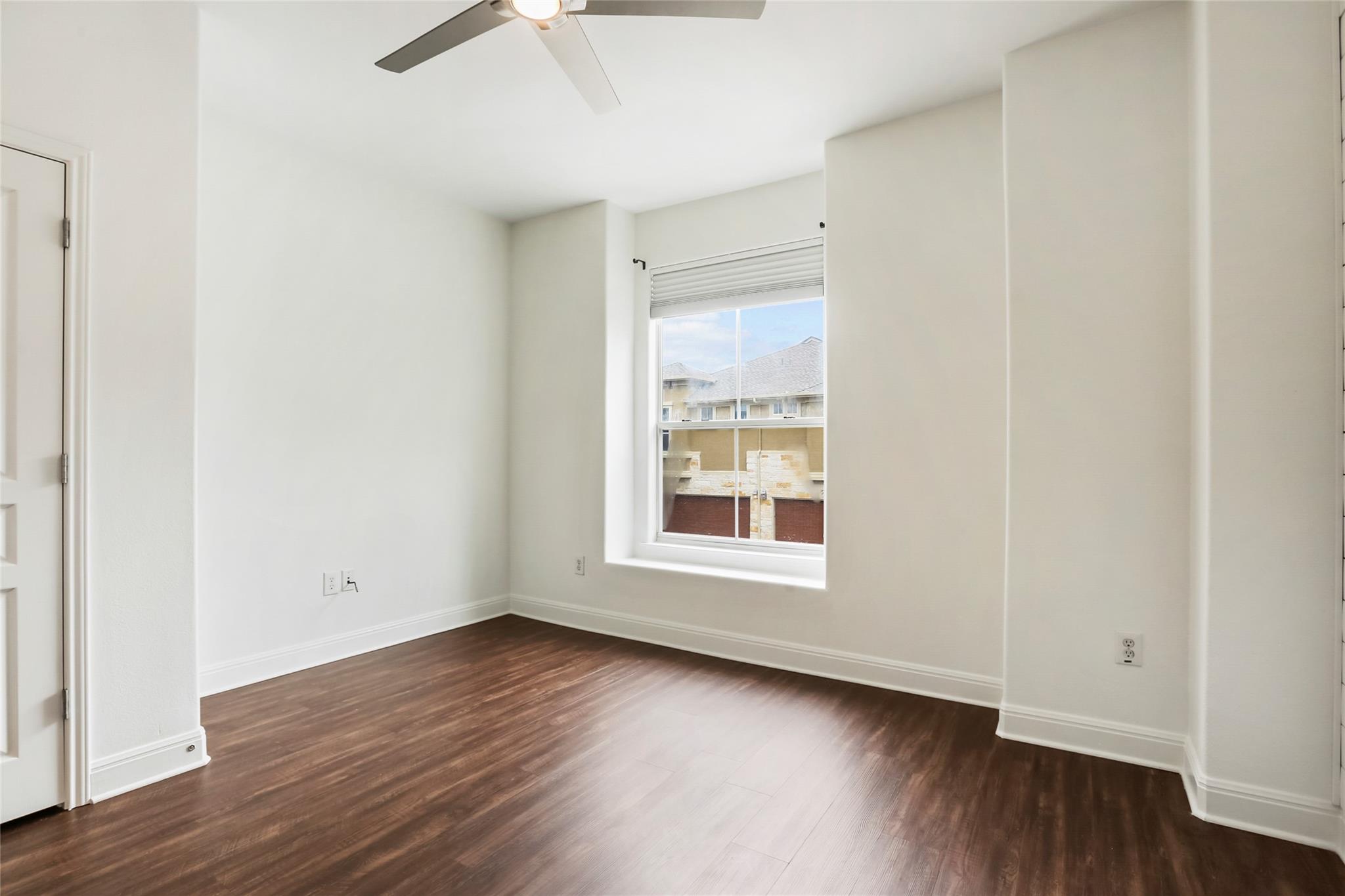 Empty room with dark wood flooring, a ceiling fan, and a window overlooking a neighboring building.