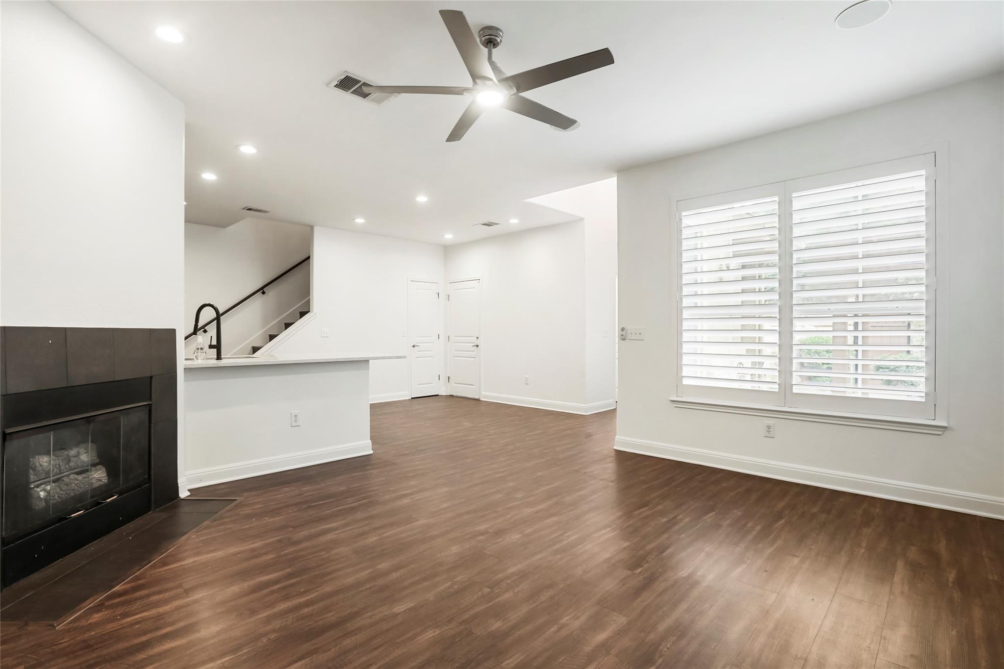 Vacant living area with dark wood floors, a black fireplace, staircase, and large shuttered windows.