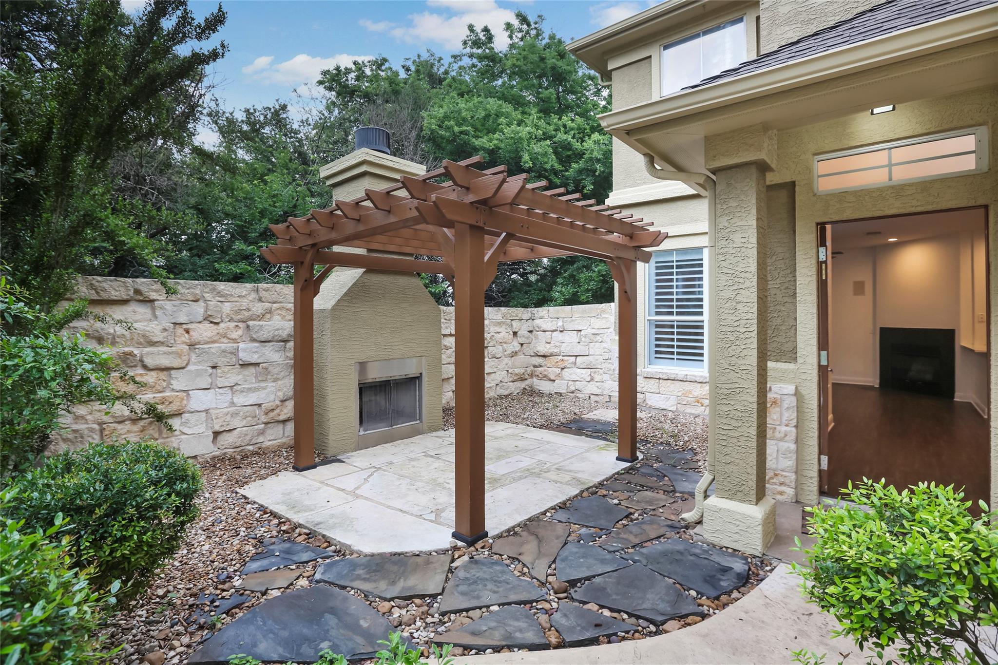 Stone courtyard with a wooden pergola, outdoor fireplace, and open doorway to the home.