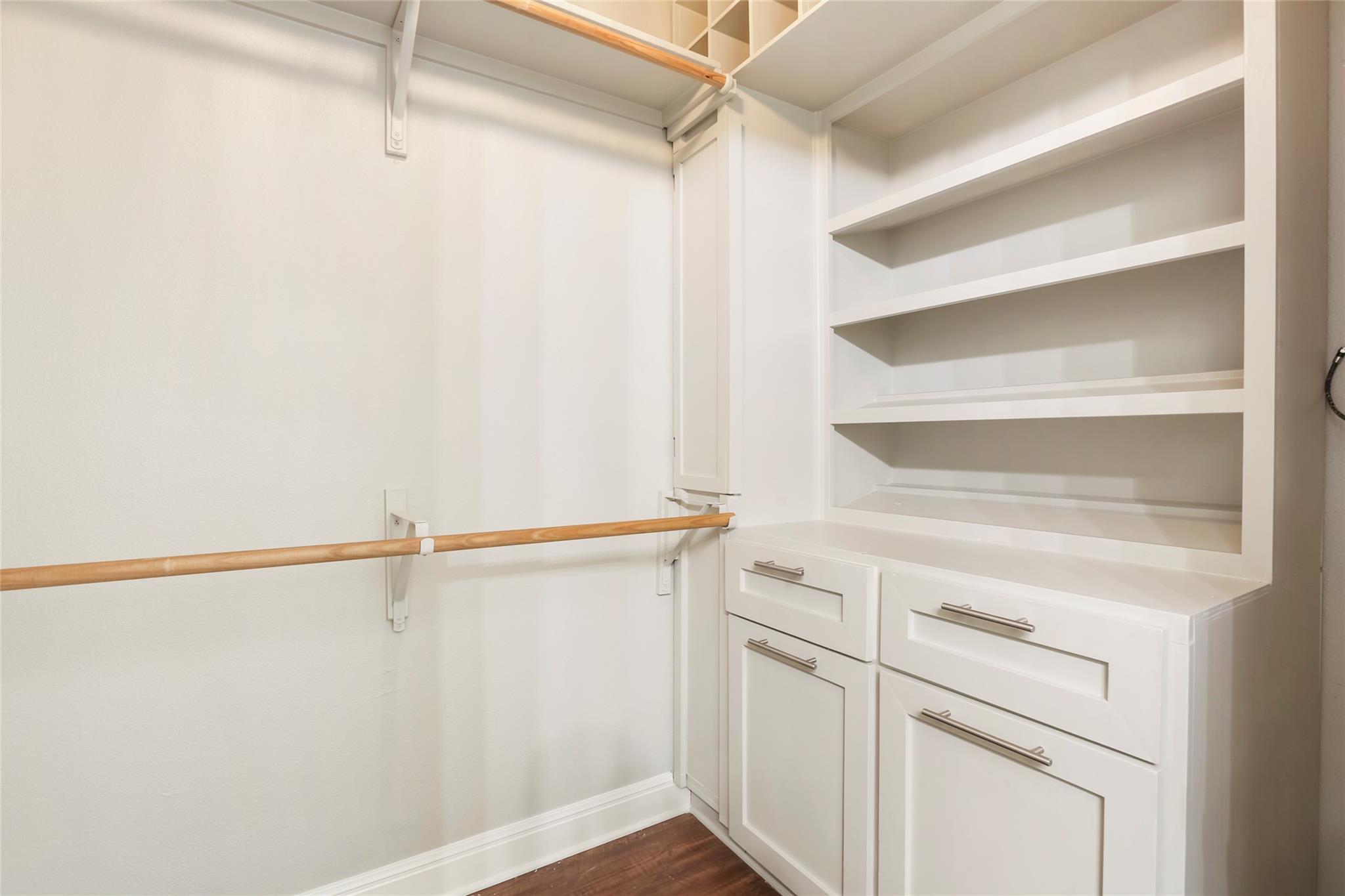 Walk-in closet with built-in white shelving and drawers, wooden hanging rods, and dark wood flooring.