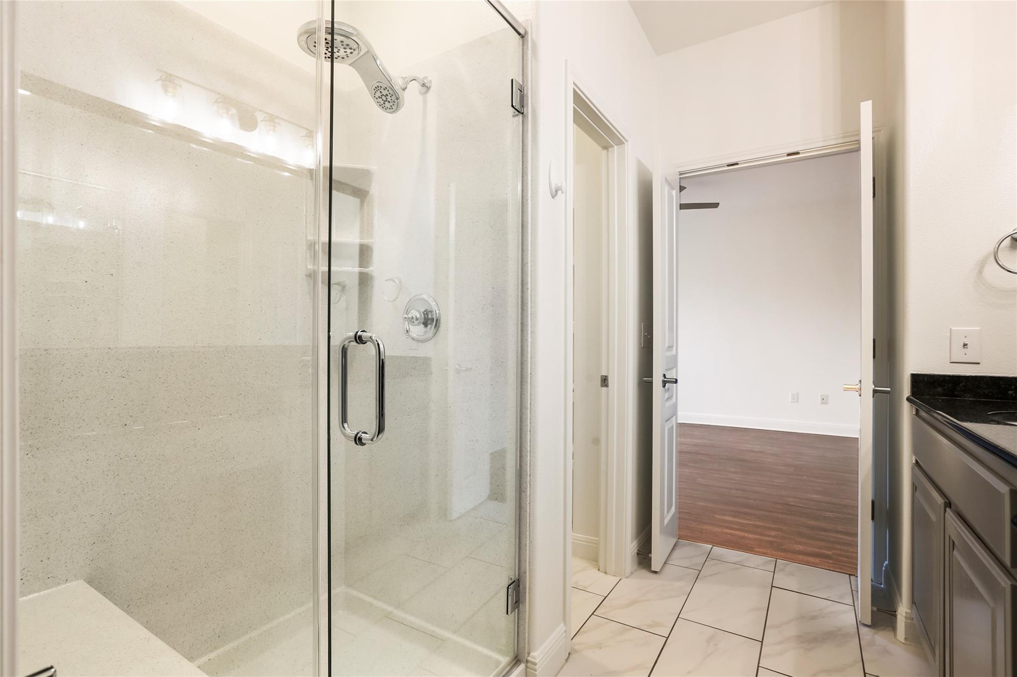 Bathroom with a glass-enclosed shower and dark vanity beside an open doorway to a room with wood-look flooring.