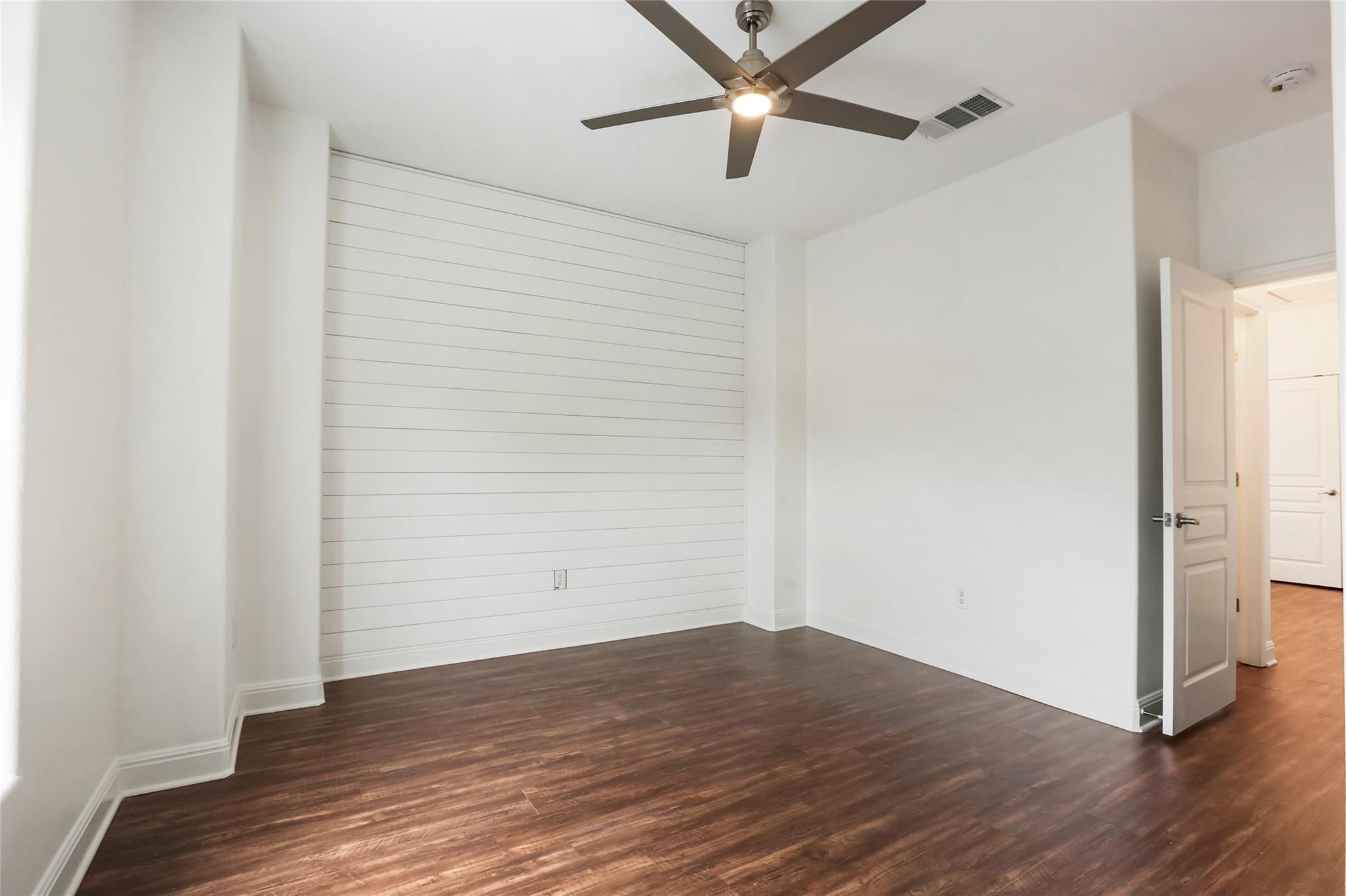 Empty bedroom with dark wood floors, a white plank accent wall, and a ceiling fan.