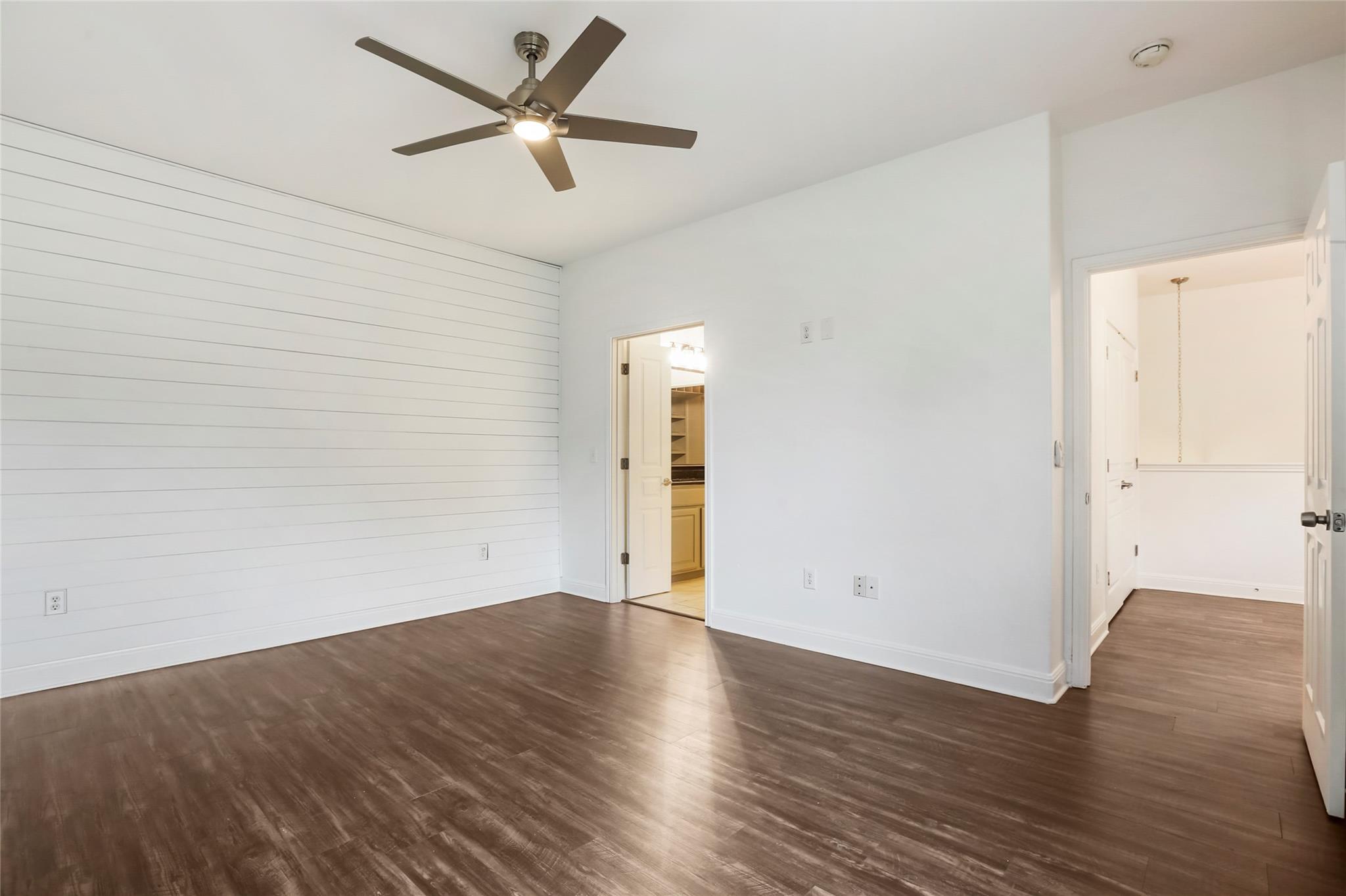 Empty bedroom with a shiplap accent wall, dark flooring, and ceiling fan; bathroom doorway at right.