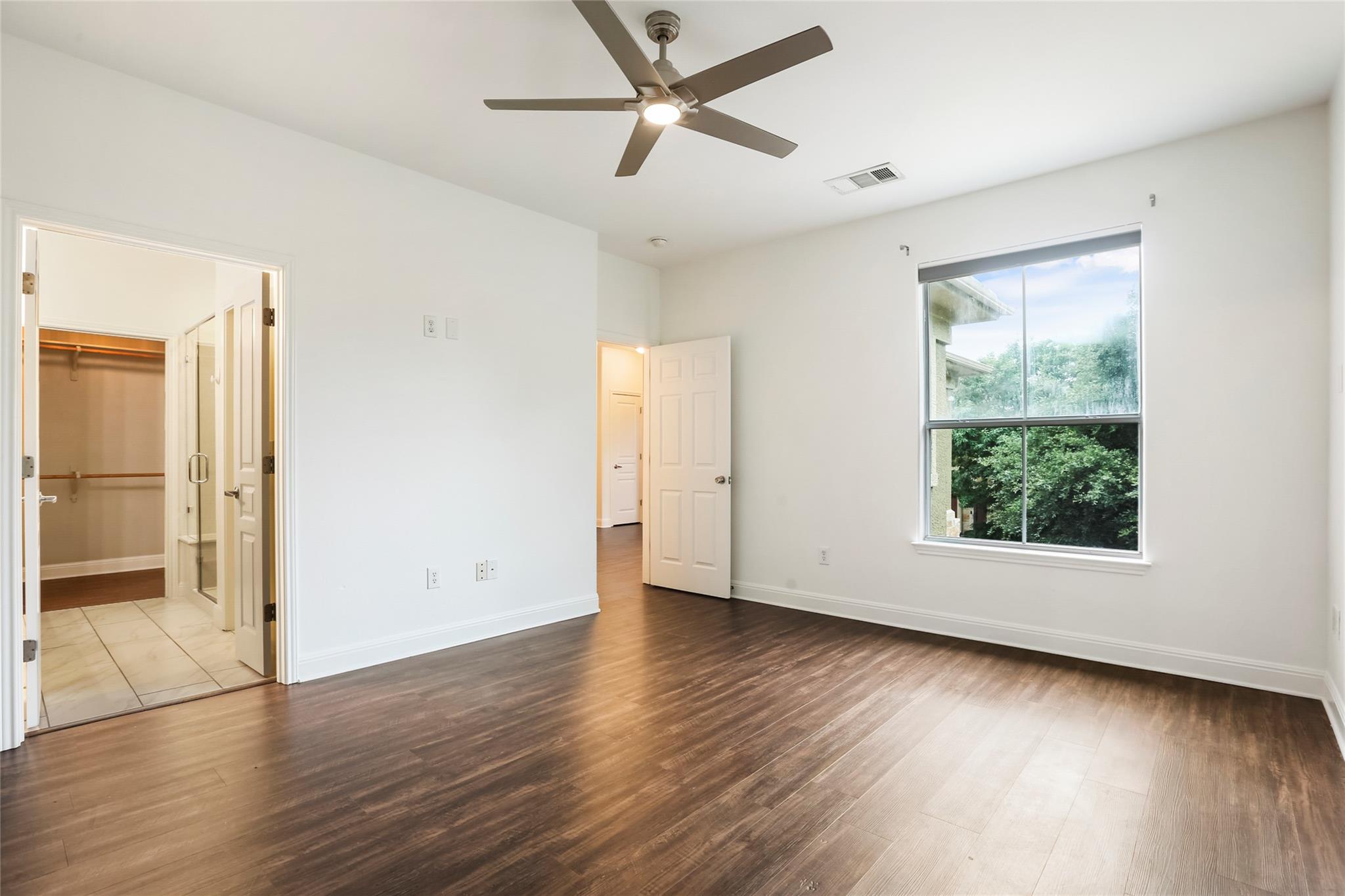 Empty bedroom with dark flooring, ceiling fan, large window, and adjoining bathroom and closet.
