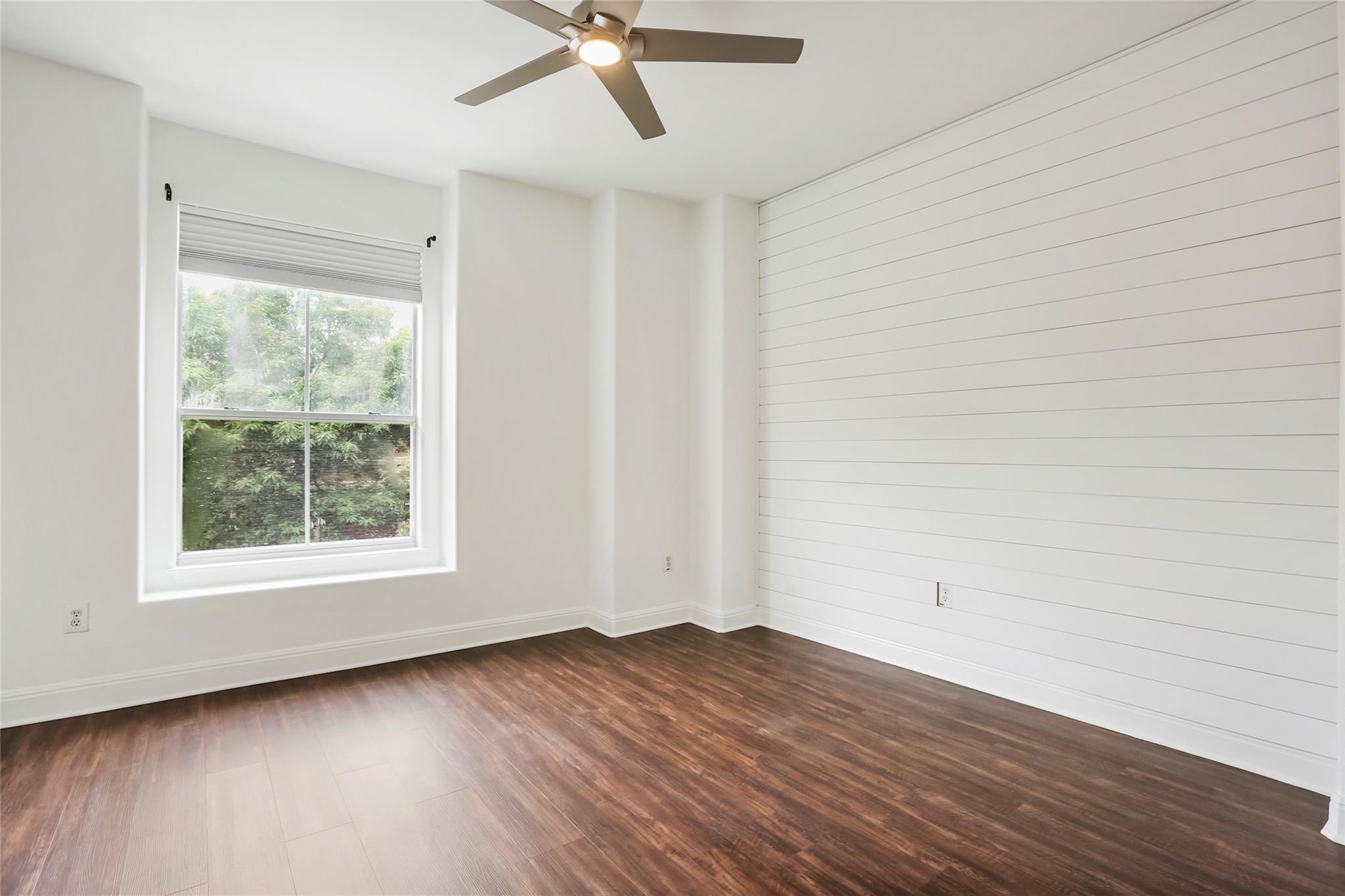 Vacant room with a white plank accent wall, dark flooring, a large window, and a ceiling fan.