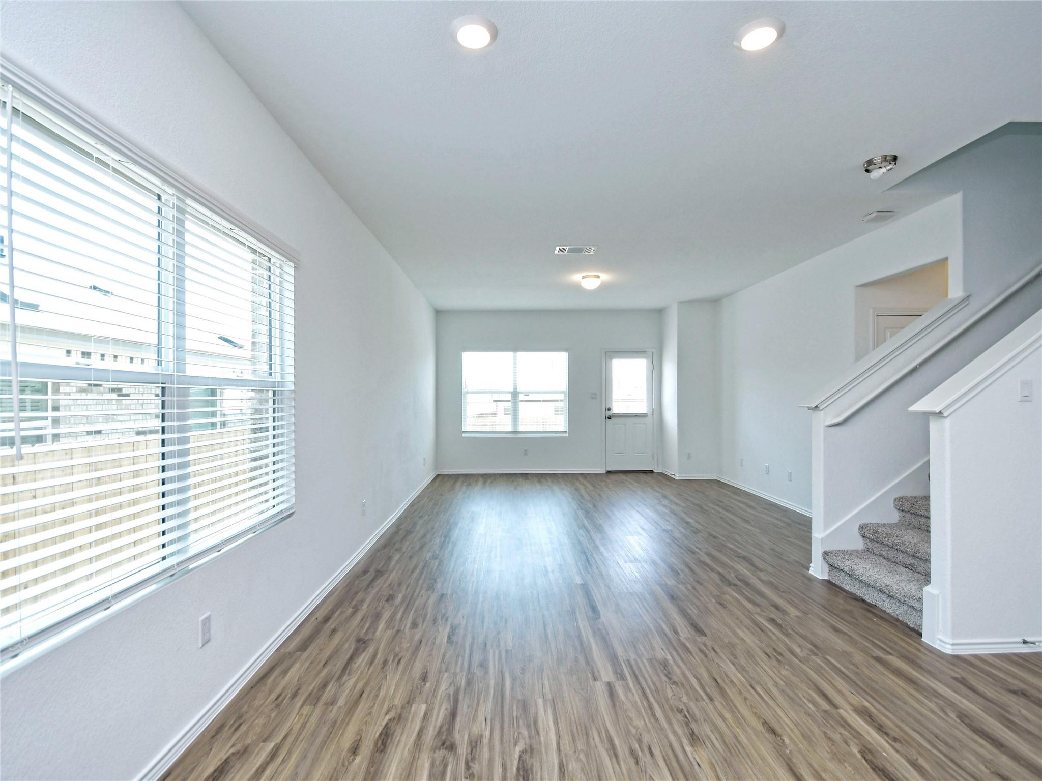 Vacant living room with wood-look floors, a large window with blinds, and carpeted stairs.