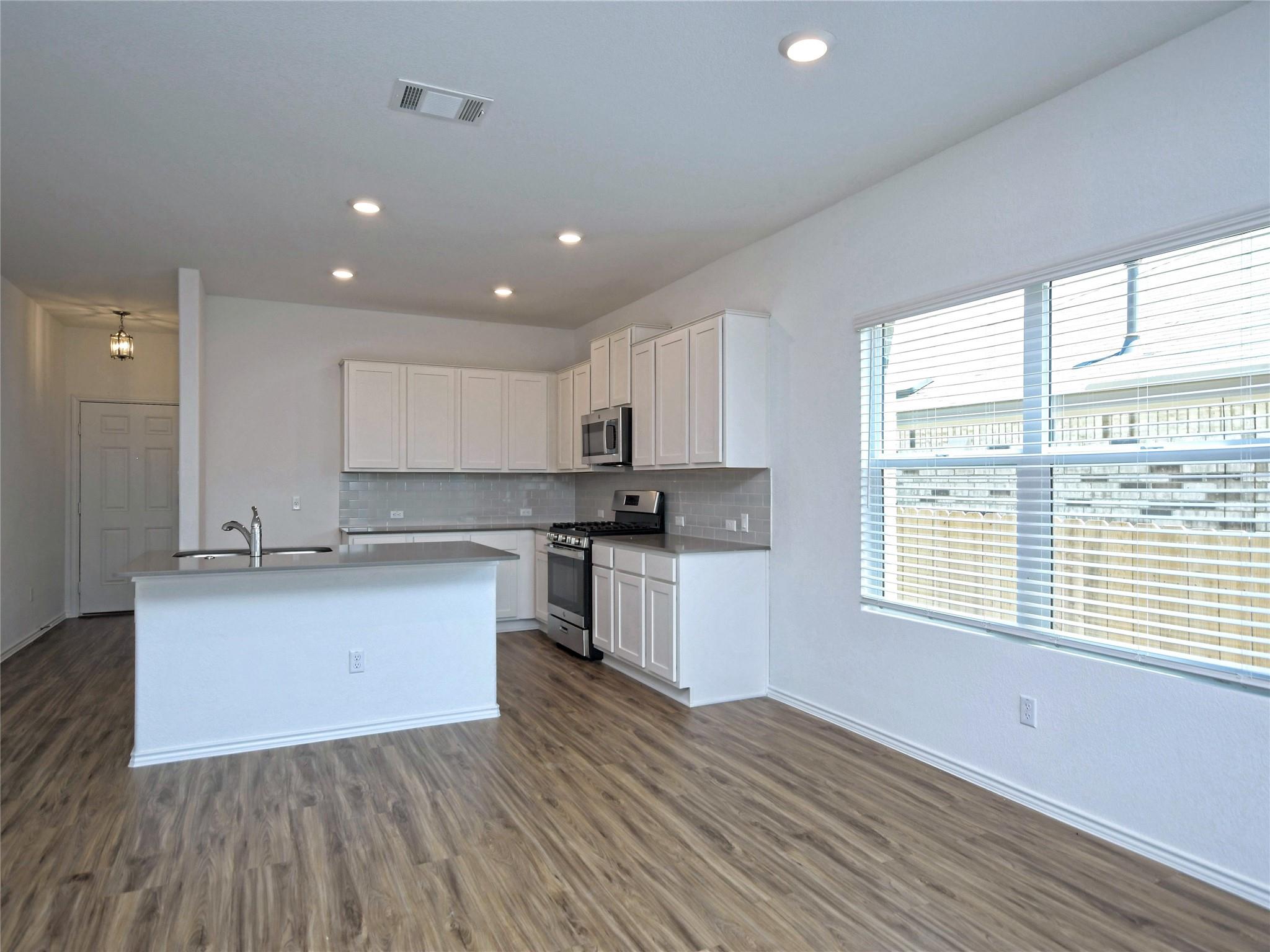 Vacant kitchen with white cabinets, stainless appliances, peninsula sink, and large window with blinds.