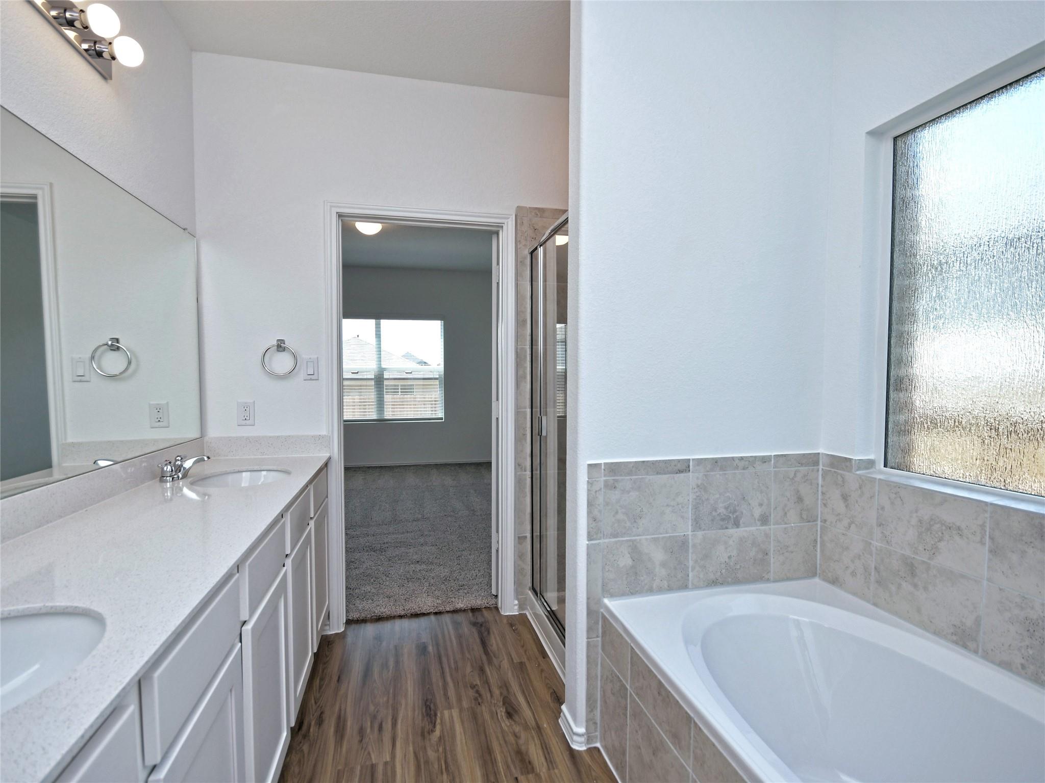 Bathroom with a long white vanity, tiled bathtub, textured privacy window, and wood-look flooring.